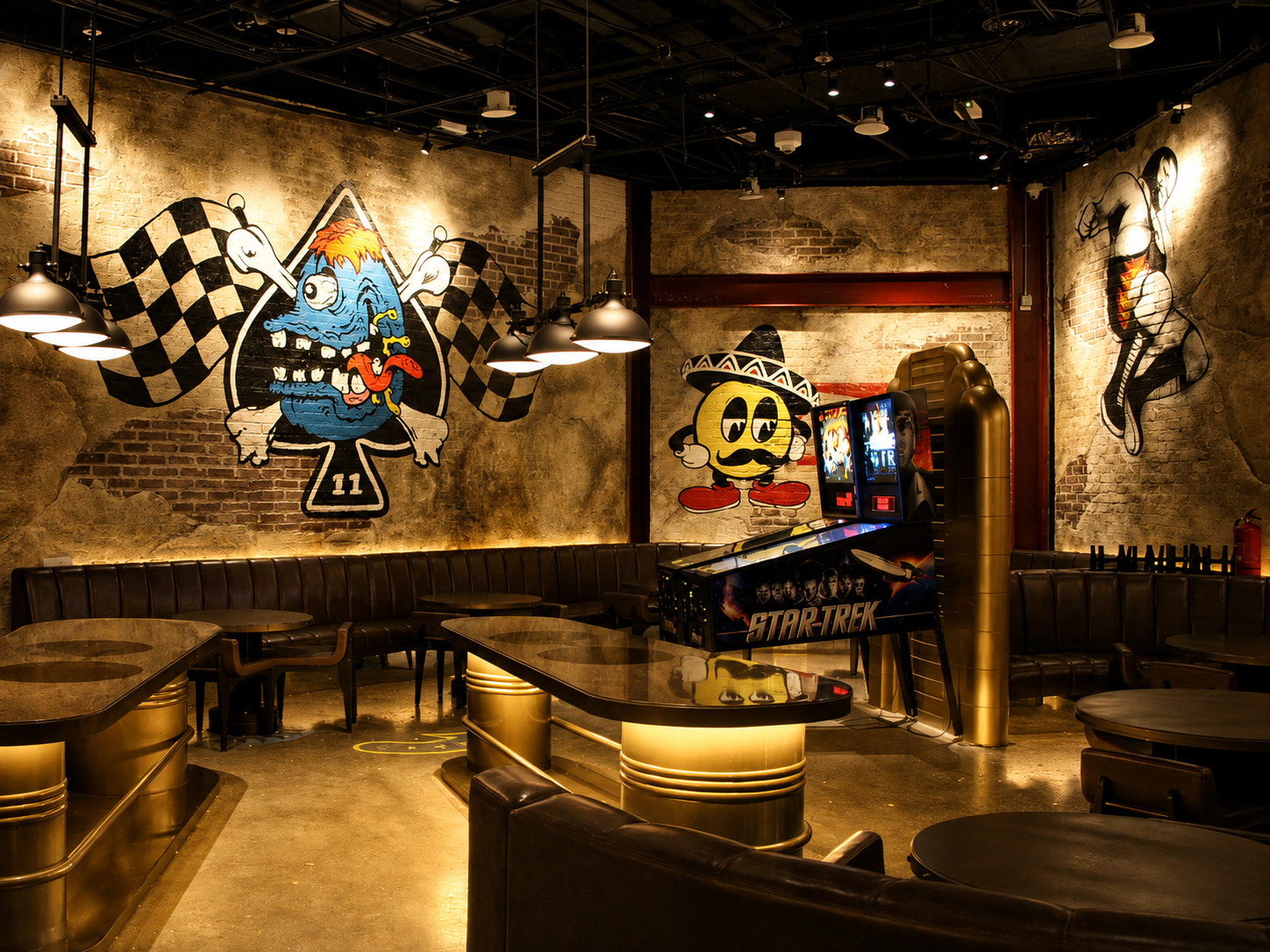 A dimly lit arcade room with brick walls and wall art including a colorful cartoon fish with checkered wings, a cartoon character wearing a sombrero, and a ballet dancer. There are tables, chairs, a pinball machine, and arcade game machines with a Star Trek theme.