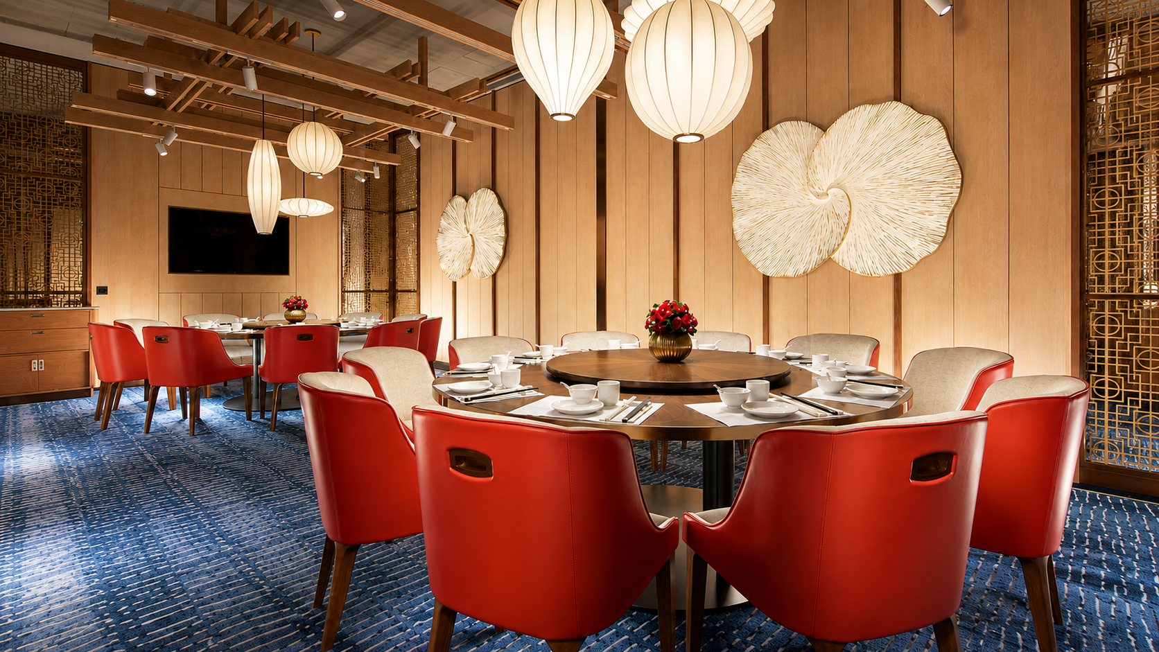 A elegantly decorated restaurant dining room with a large round table set for a meal. The table is surrounded by red and beige chairs, with a blue patterned carpet on the floor. The walls are wood-paneled with decorative wall art, and there are hanging lantern-style light fixtures from the ceiling.