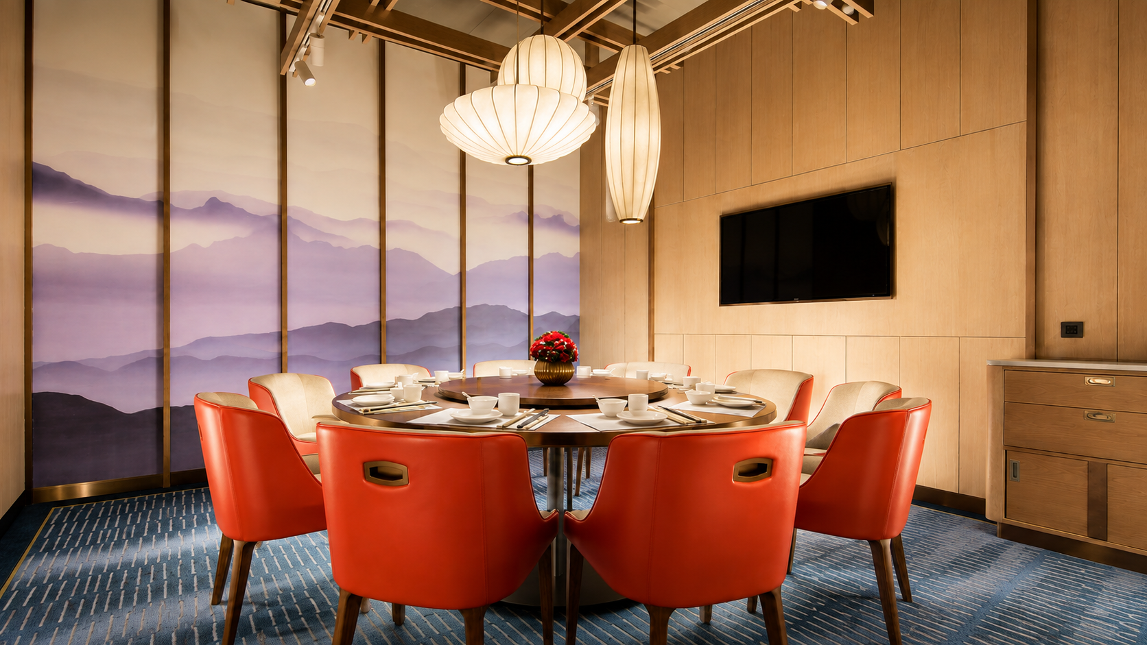 Elegant dining room with a round table set with white dishes and chopsticks, surrounded by red leather chairs. A floral centerpiece decorates the table. The room features a wall with purple mountain mural, a mounted TV, and warm wooden walls with hanging paper lantern and pendant lights.