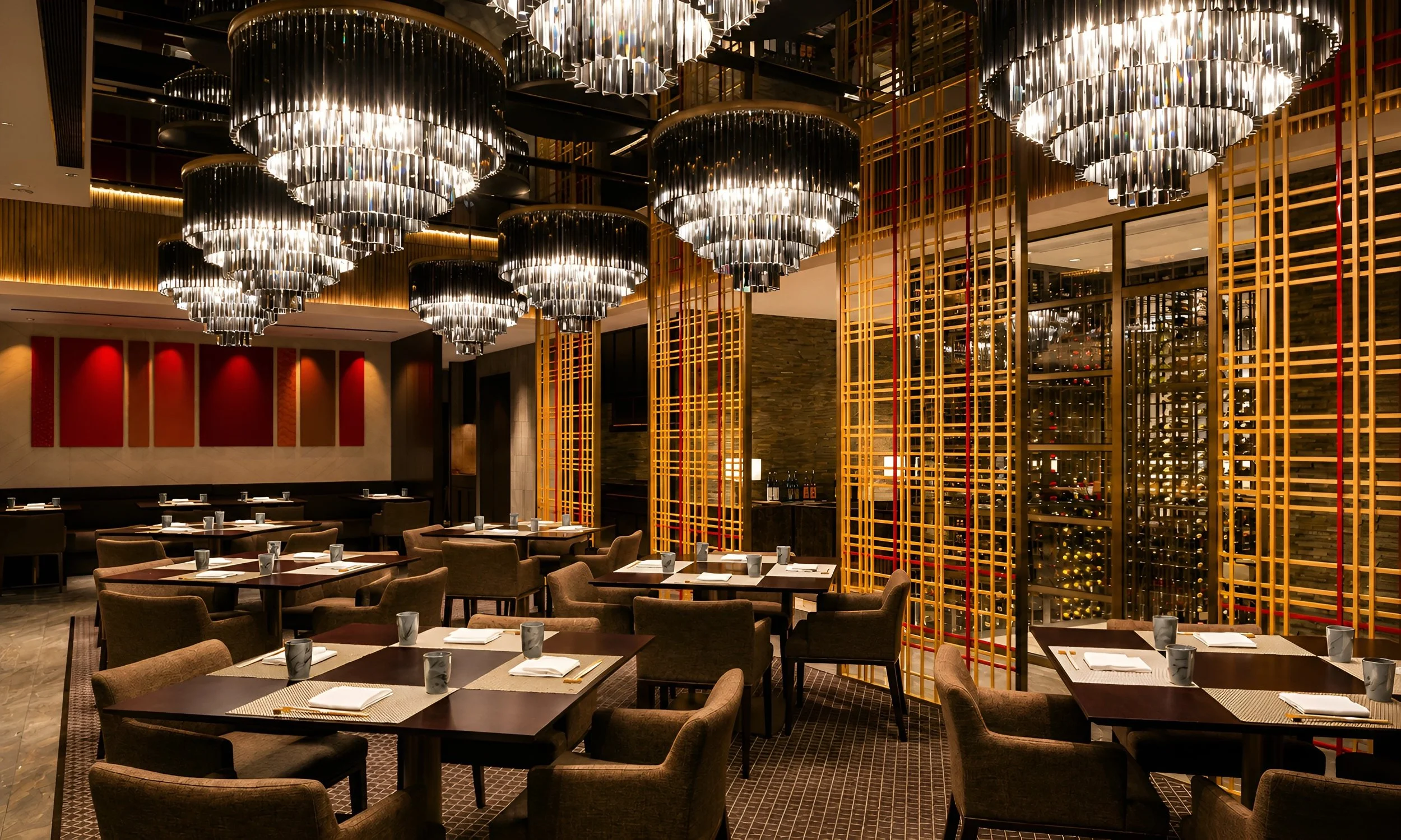 Interior of a restaurant with multiple tables set with placemats, napkins, and cups, and modern chandeliers hanging from the ceiling. The decor includes wooden wall panels and a large window with wine bottles displayed behind decorative wooden slats.
