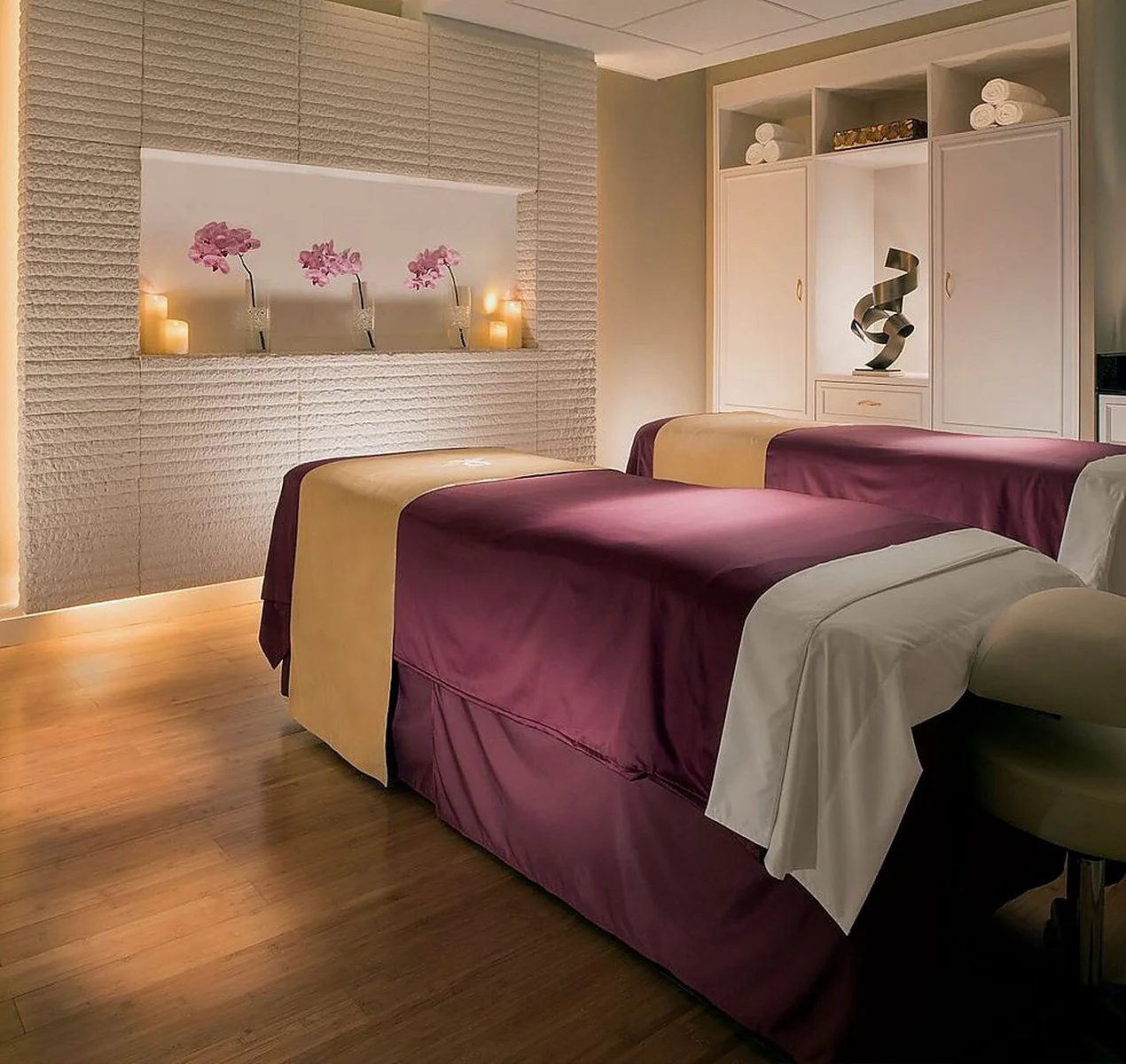 A serene massage room with two massage tables covered in purple and beige sheets. The room features bamboo flooring, a textured wall with a built-in niche holding pink flowers and lit candles, and a large white cabinet with rolled towels and a decorative sculpture.