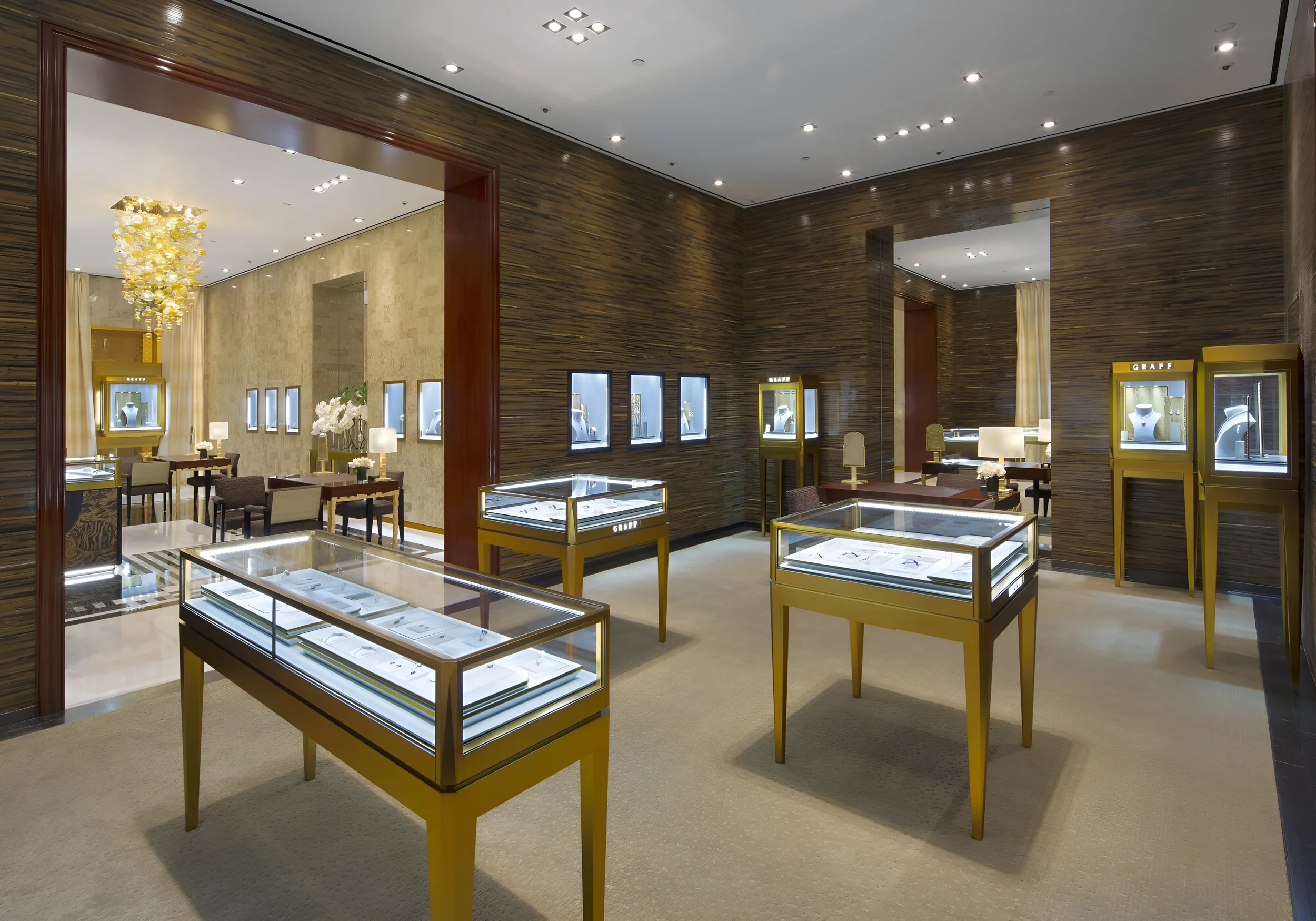 Interior of a jewelry store with display cases showcasing jewelry, gold accents, seating area, modern chandelier, wood-paneled walls, ceiling lighting, and framed artwork on walls.