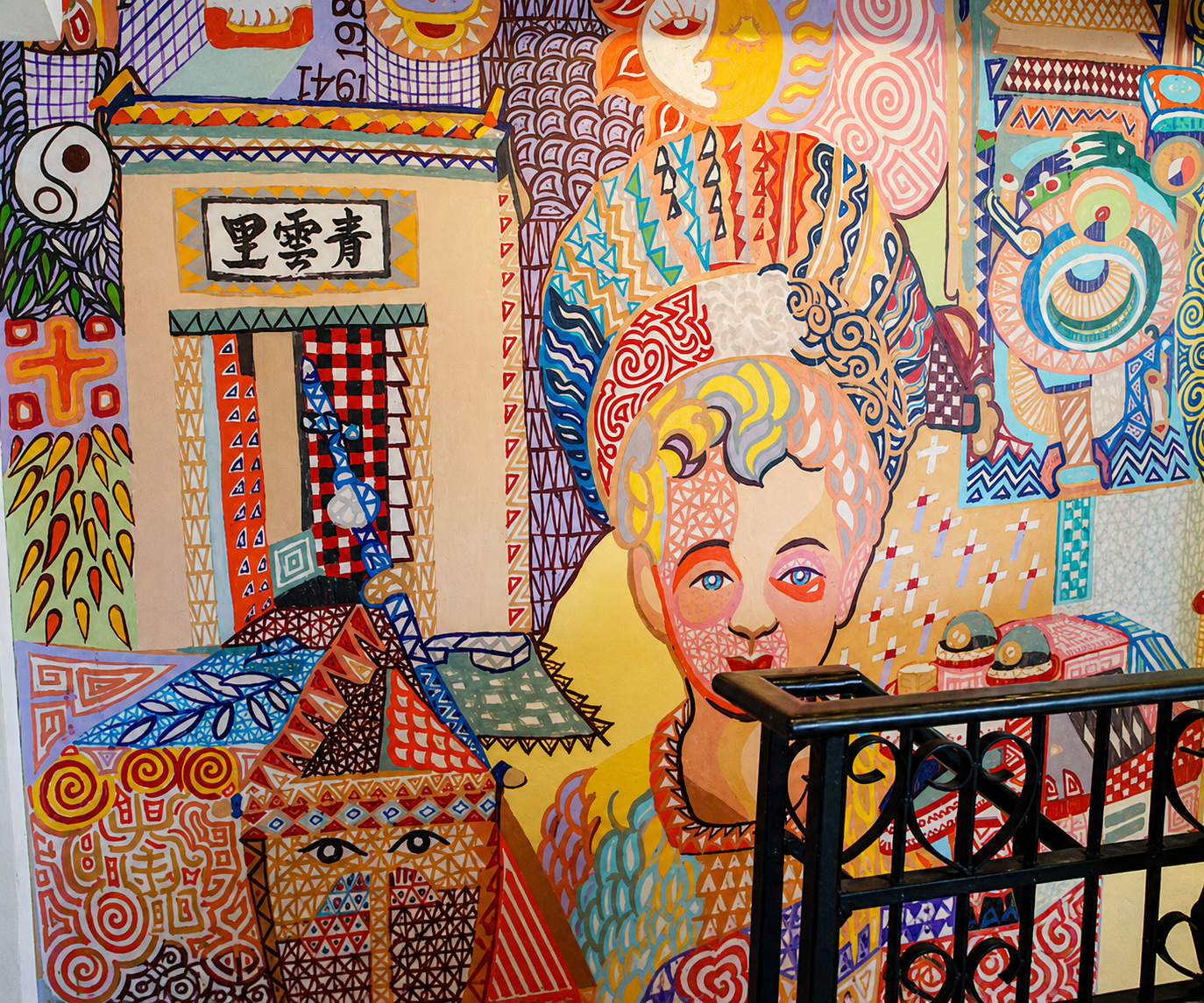 Colorful mural with abstract faces, sun, moon, and various geometric patterns. It features a large female face with blonde hair, surrounded by vibrant shapes and symbols. There is a black railing in front of the mural.