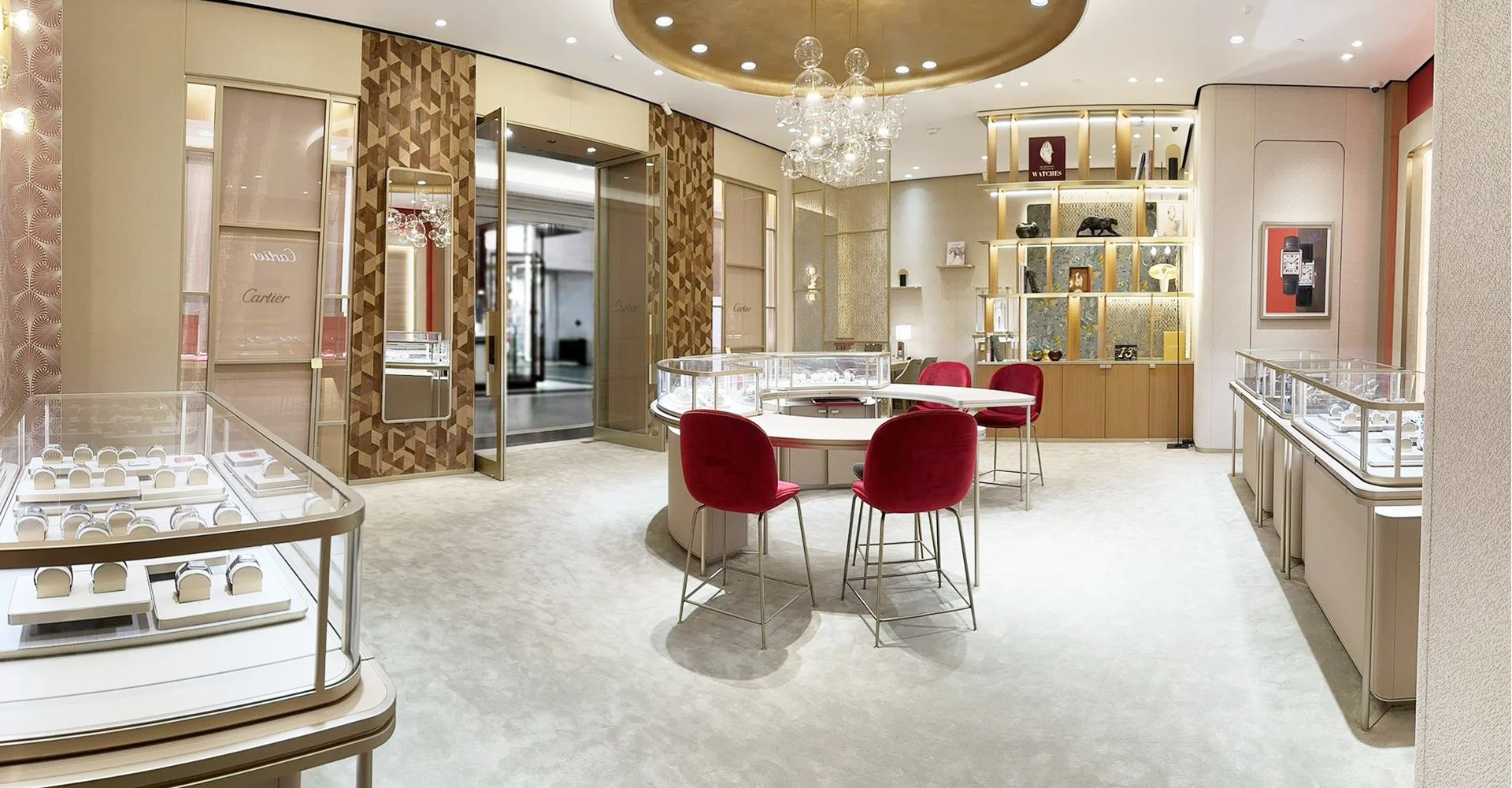 Interior of a jewelry store with display cases filled with jewelry, a small round table with red chairs, decorative shelving with art pieces, and a chandelier.