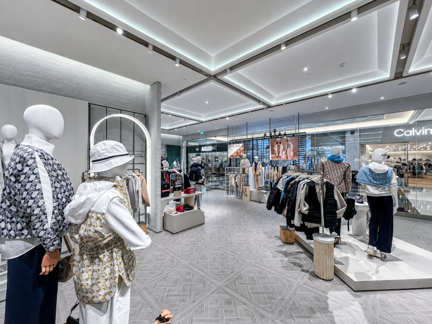 Inside a modern clothing store, mannequins dressed in casual outfits display on the left and right, with various clothing racks and displays in the background. The store has high ceilings, recessed and track lighting, and a glass storefront.