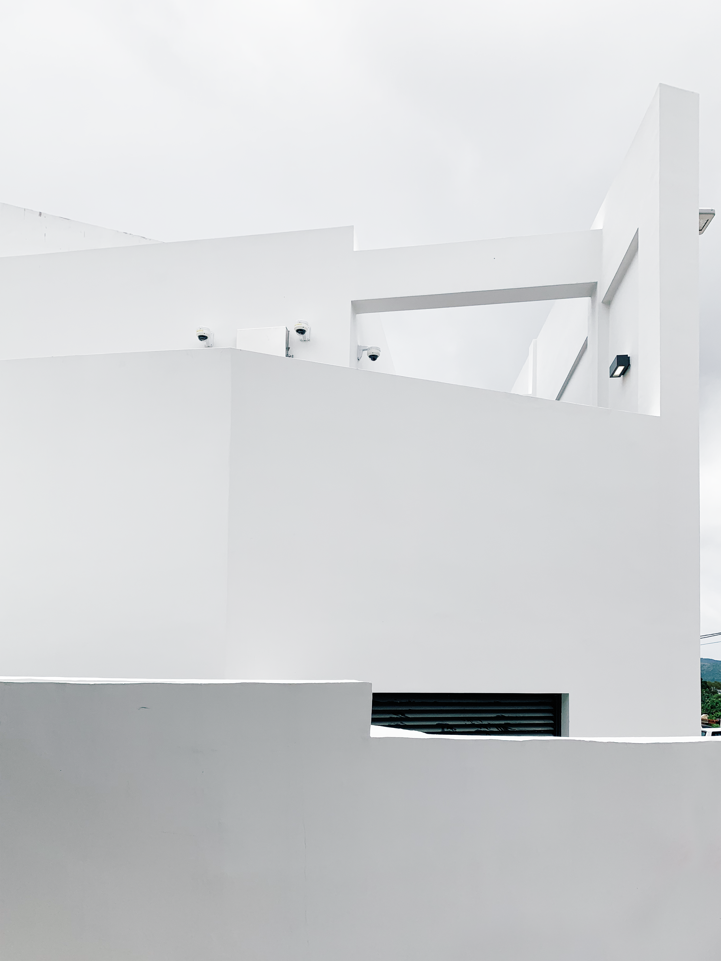 Modern white building with geometric design, security cameras, and external lighting fixtures under an overcast sky.
