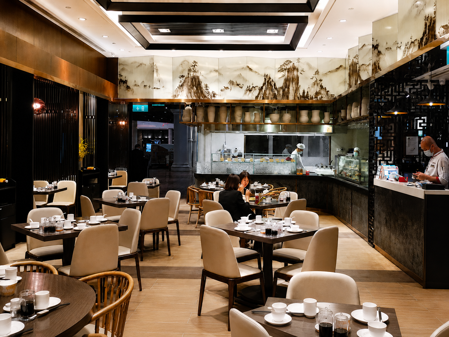 Inside a modern restaurant with beige chairs, dark wooden tables set with cups and glasses, and a kitchen area with chefs preparing food. Decor includes traditional Asian art and pottery.