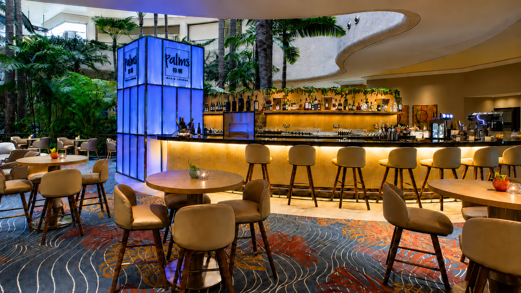 Indoor bar area with a curved bar counter, beige bar stools, round tables with chairs, lush green plants, and a blue-lit sign that reads 'Palms Bar & Lounge', in a tropical-themed setting.