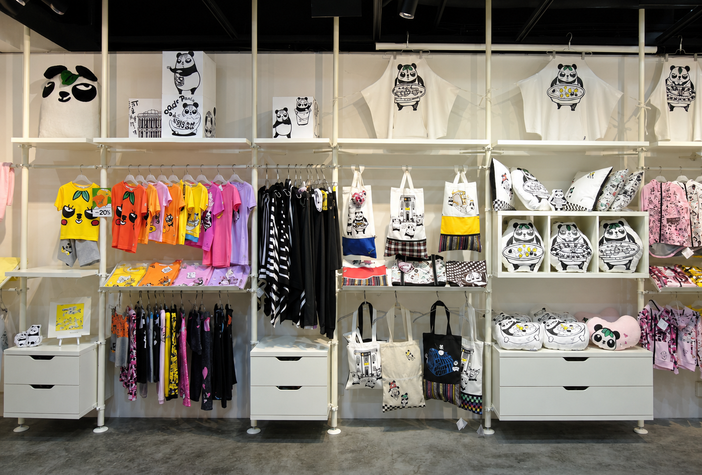 Display of panda-themed clothing and accessories in a store, including plush toys, t-shirts, tote bags, pillows, and clothing with panda illustrations in black, white, and colorful designs.