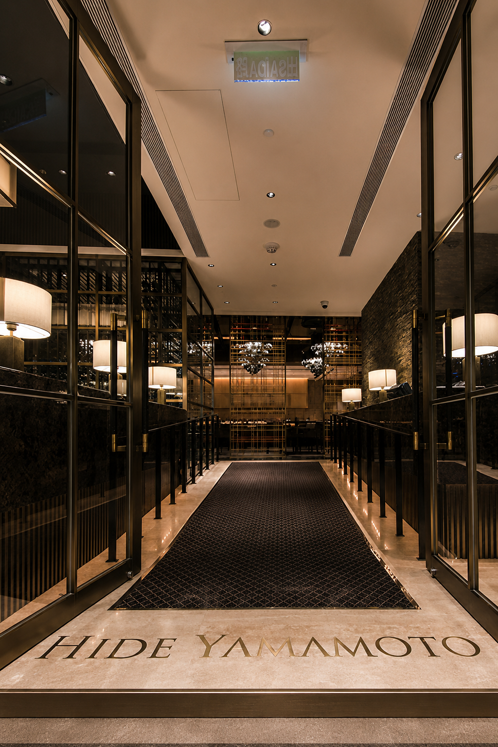 Elegant hotel lobby entrance with black glass doors, framed by gold accents, leading to a dimly lit interior with modern lighting and decor, and the words 'HIDE YAMAMOTO' on the floor mat.