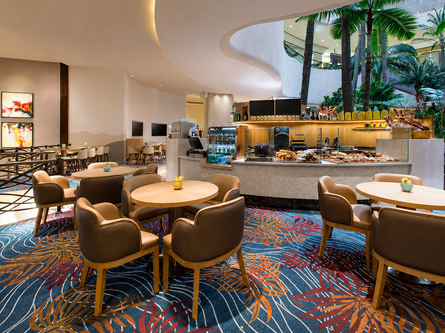 Interior view of a modern hotel lobby with a breakfast area, featuring round tables with beige chairs, a food counter with baked goods, tropical plants, and a colorful, palm leaf patterned carpet.