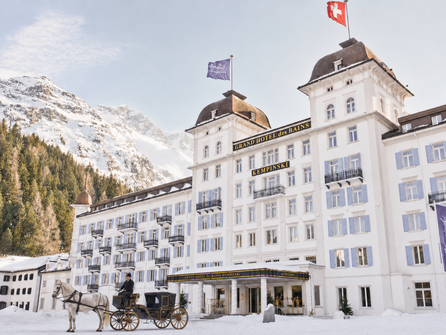 Das Bild zeigt das Grand Hotel des Bains Kempinski in einer verschneiten Berglandschaft. Vor dem Hotel ist eine Kutsche mit einem Pferd und einem Kutscher im Outfit eines Jahrhunderts. Das Hotel ist weiß mit blauen Fensterläden und hat mehrere Balkone. Hinter dem Hotel sind hohe, schneebedeckte Berge und ein bewölkter Himmel.