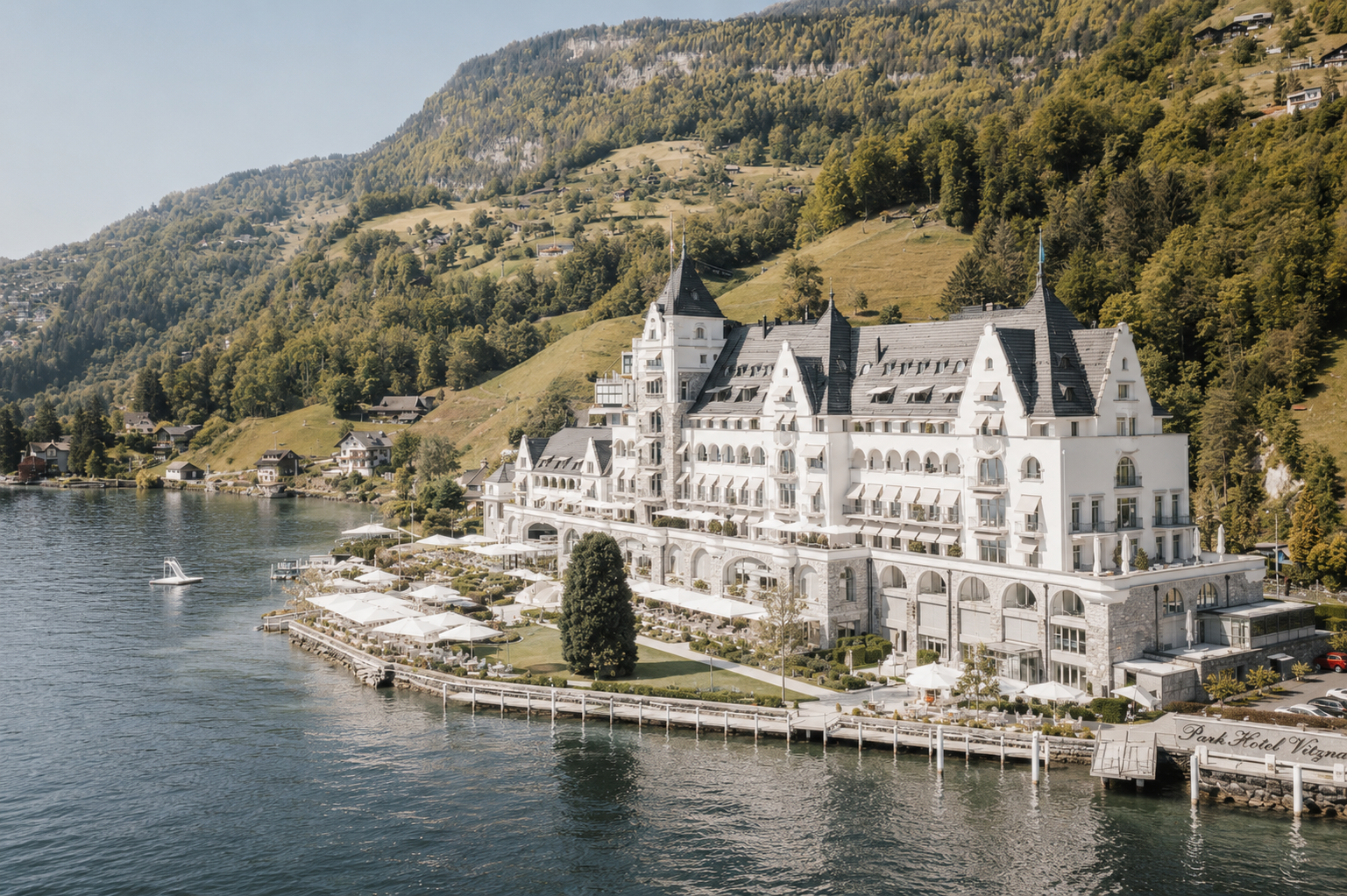 Luxushotel am See mit weißen Fassaden, Terrassen und Sonnenschirmen, umgeben von Bergen und Wald, mit Wasser und Booten im Vordergrund.