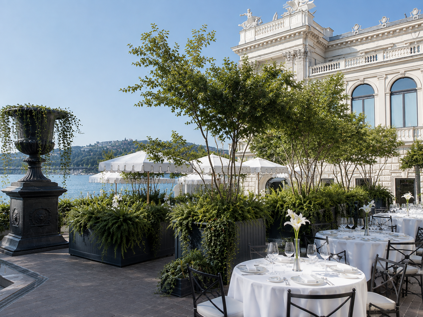 Ein eleganter Außenbereich mit runden Tischen, die mit weißen Tischdecken, Gläsern und Blumenarrangements gedeckt sind, neben einer Anlegestelle am Wasser, vor einem weißen historischen Gebäude mit großen Fenstern und Verzierungen. Es gibt auch weiße Sonnenschirme und eine Uferpromenade, umgeben von Bäumen und Pflanzen.