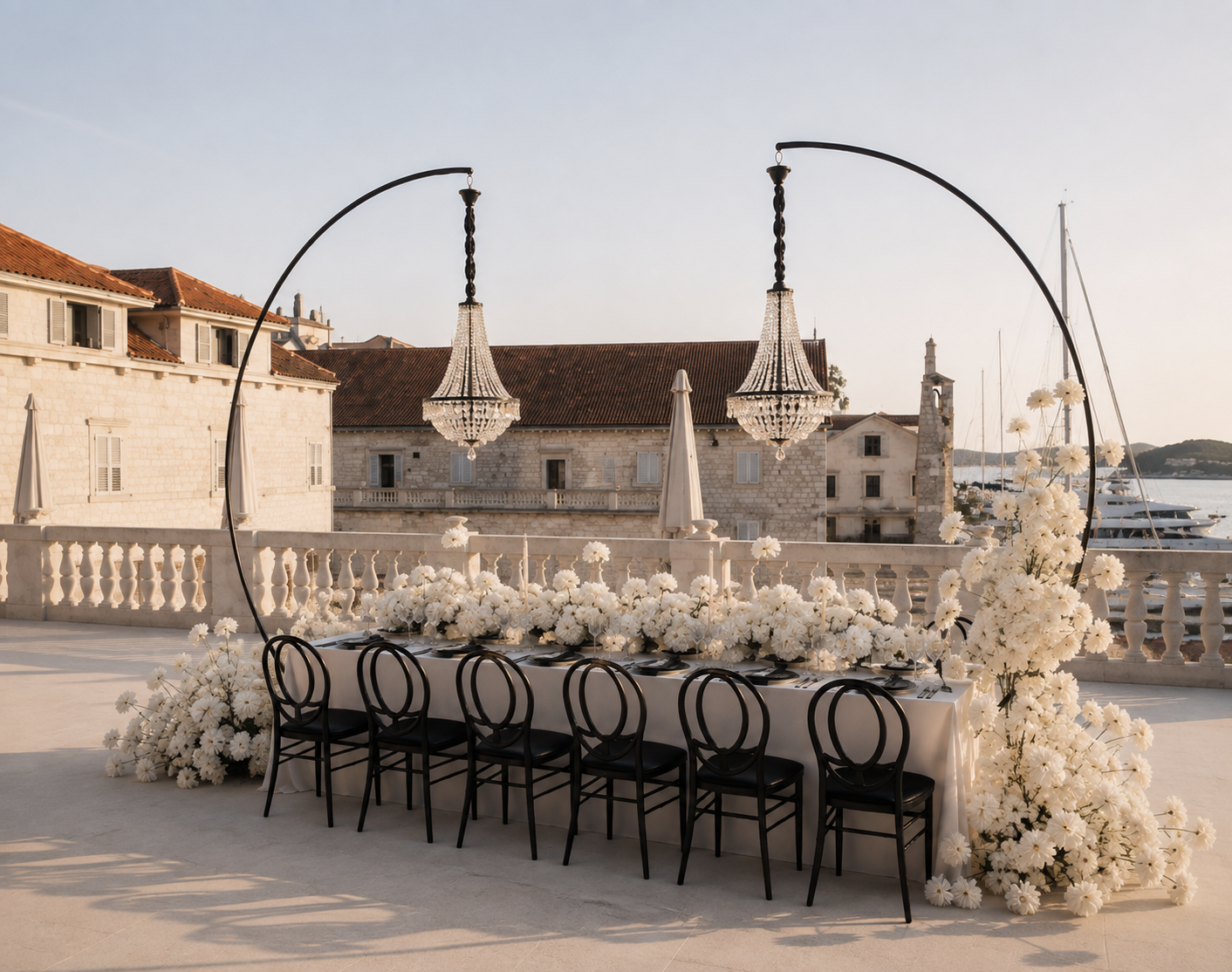 Ein eleganter, lang gedeckter Tisch mit weißen Blumen und schwarzem Geschirr, umrahmt von weißen Blumenarrangements, im Freien bei Sonnenuntergang, mit Blick auf historische Gebäude und Boote am Wasser.