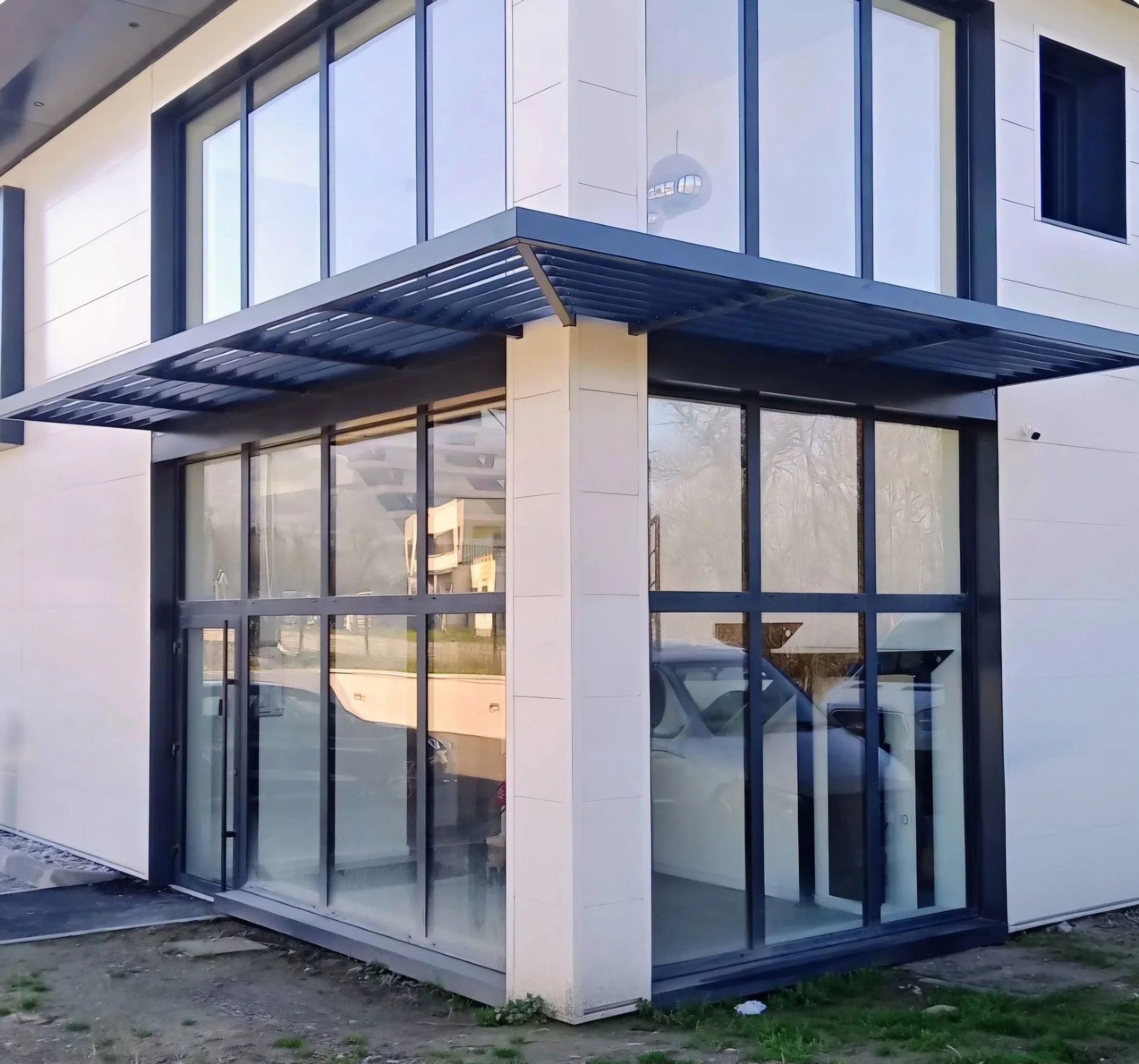 Bâtiment moderne avec grande façade en verre, structure en métal visible, et un balcon orné d'une grille métallique. La construction se trouve en extérieur, avec sol en terre et quelques herbes.