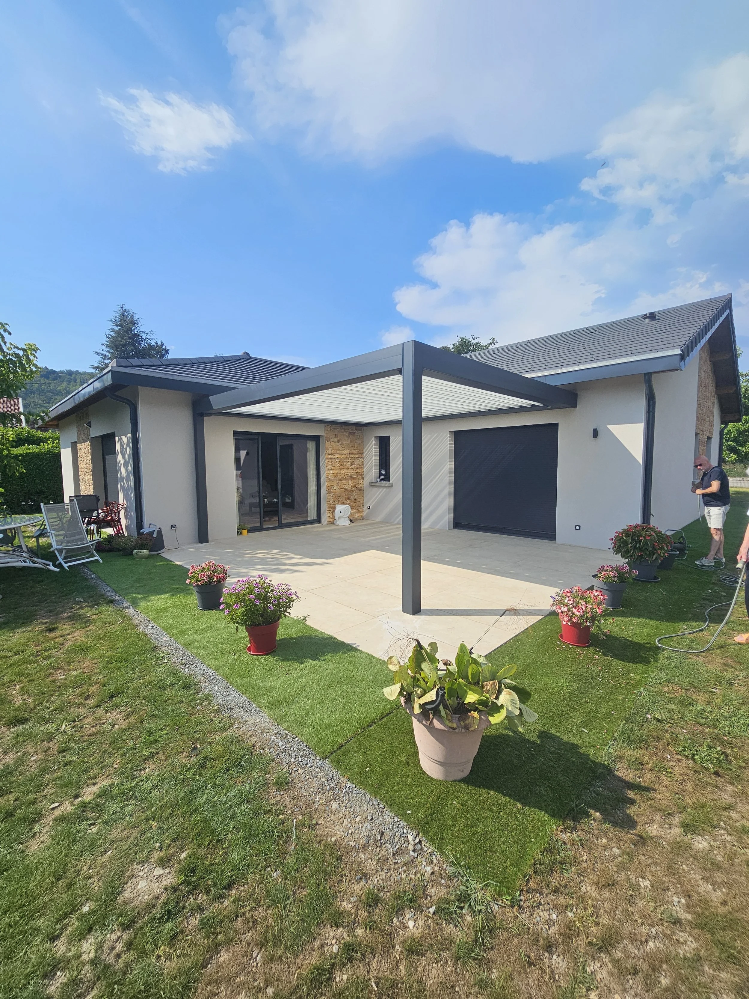 Une maison moderne avec une terrasse et un espace vert bien entretenu, des pots de fleurs colorés, et un ciel bleu avec quelques nuages.