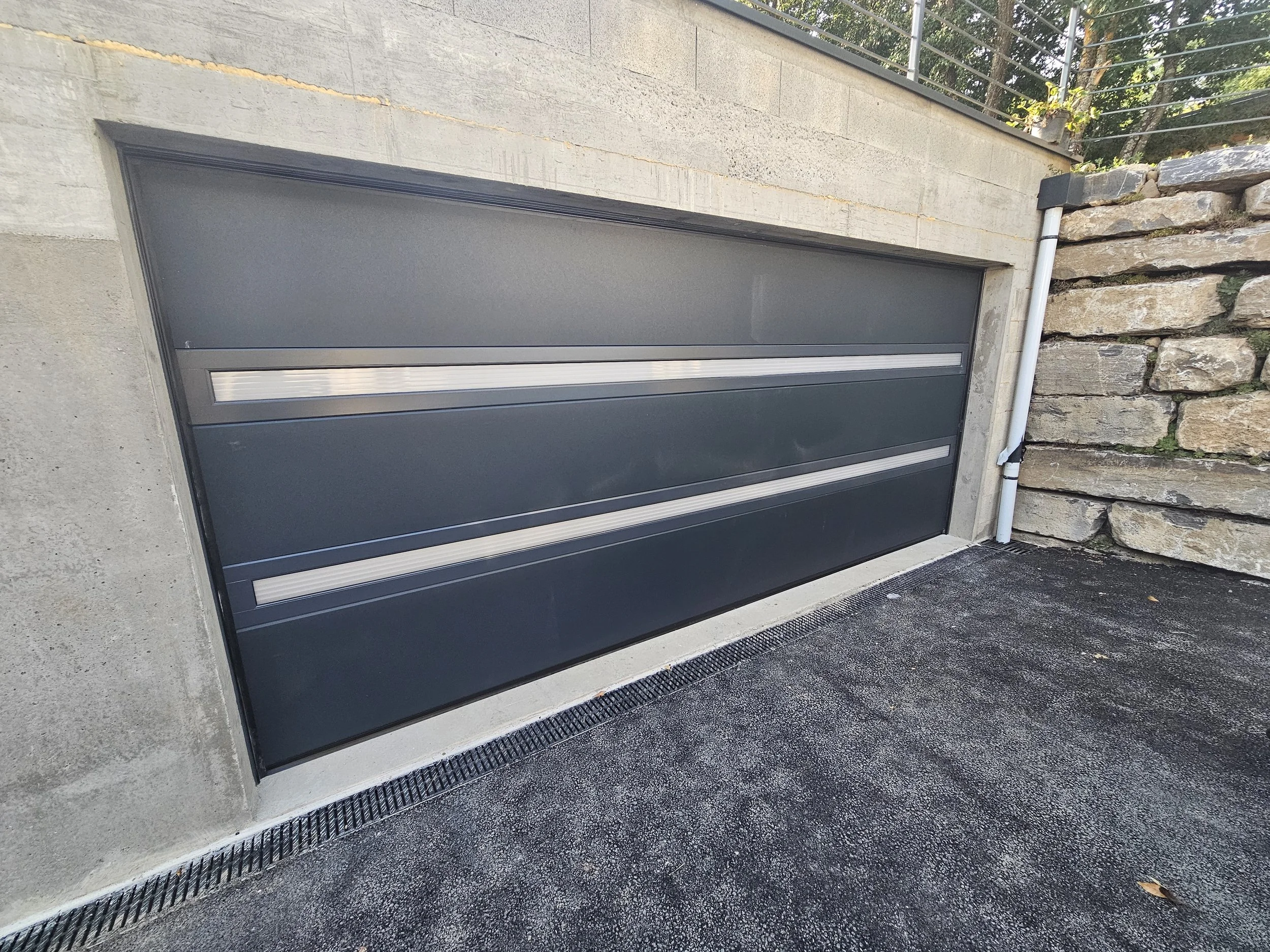 Une porte de garage gris foncé encastrée dans un mur en béton avec un revêtement en pierres à droite, un drain et un tuyau de plomberie blanc à côté.