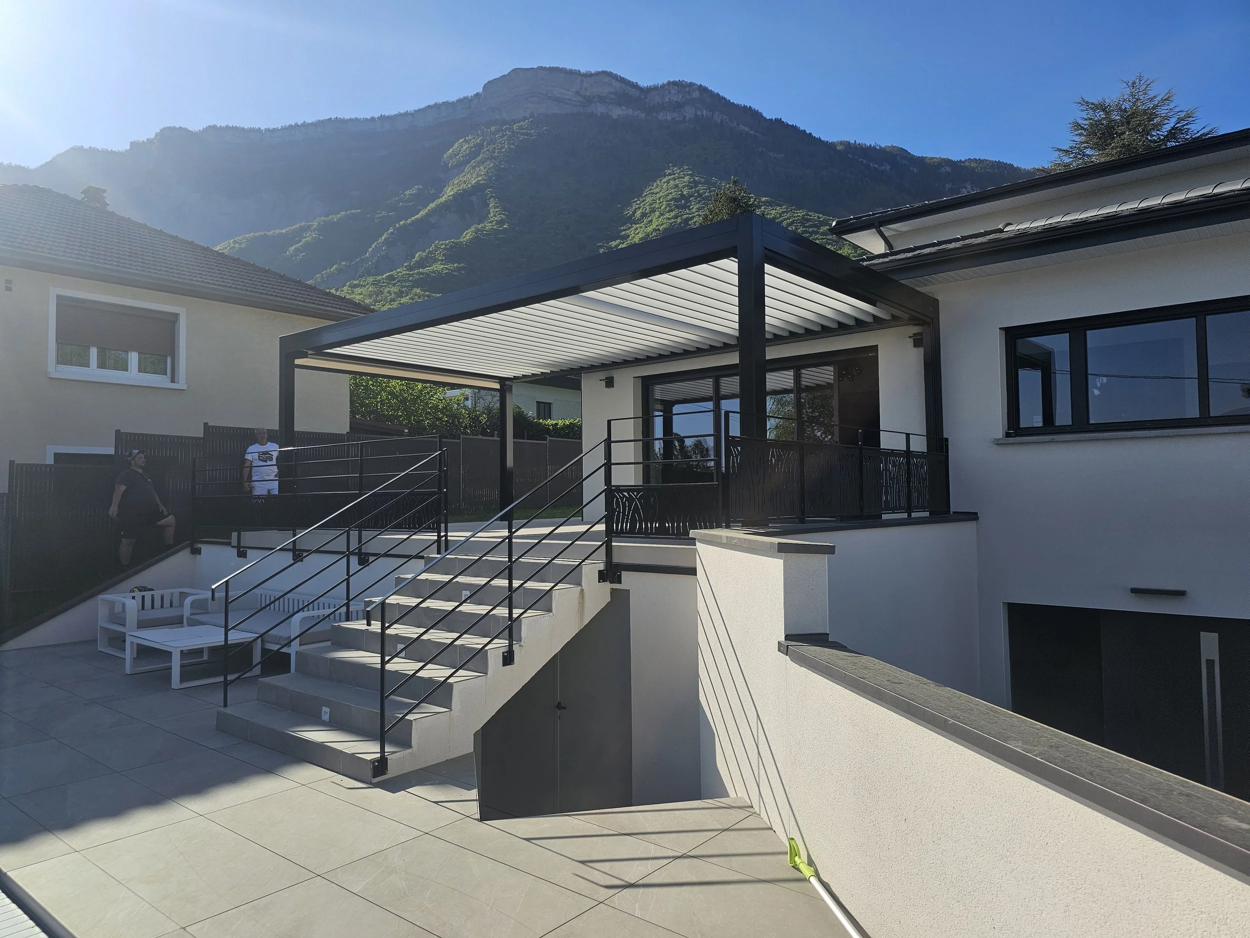 Une maison moderne avec une terrasse en extérieur, un escalier menant à un espace de vie, avec des montagnes en arrière-plan et un ciel clair.