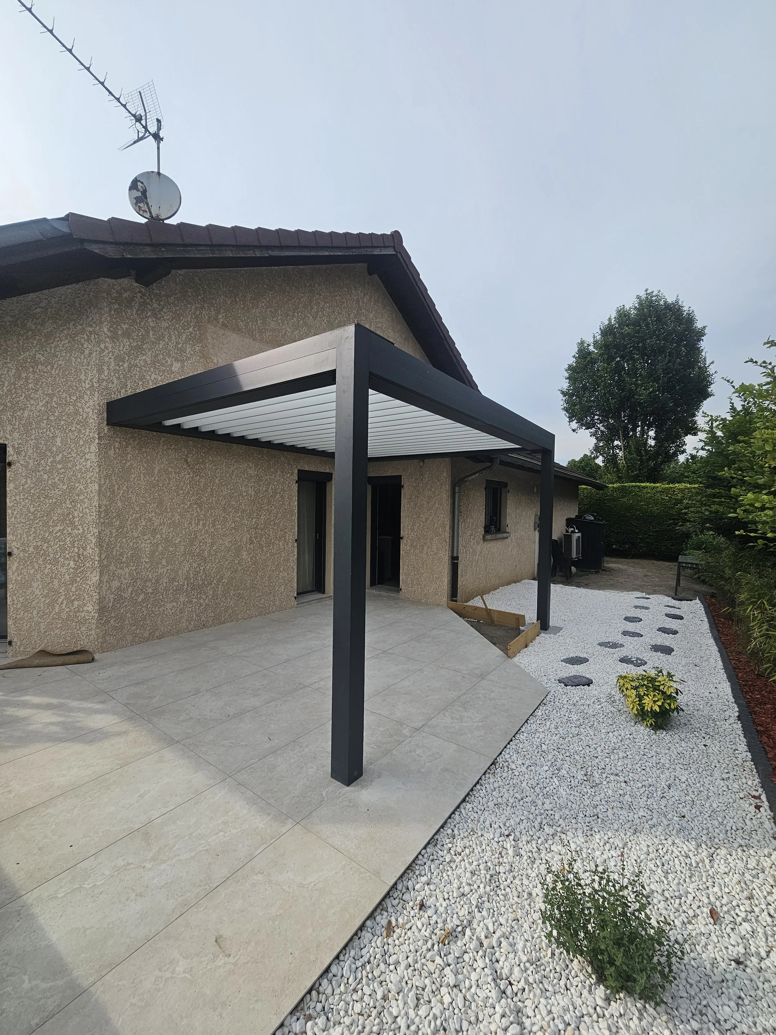 Côté extérieur d'une maison avec une terrasse carrelée recouverte d'une pergola noire, zone de gravier blanc avec plantes et un arbre en fond, et un toit en tuiles rouges.