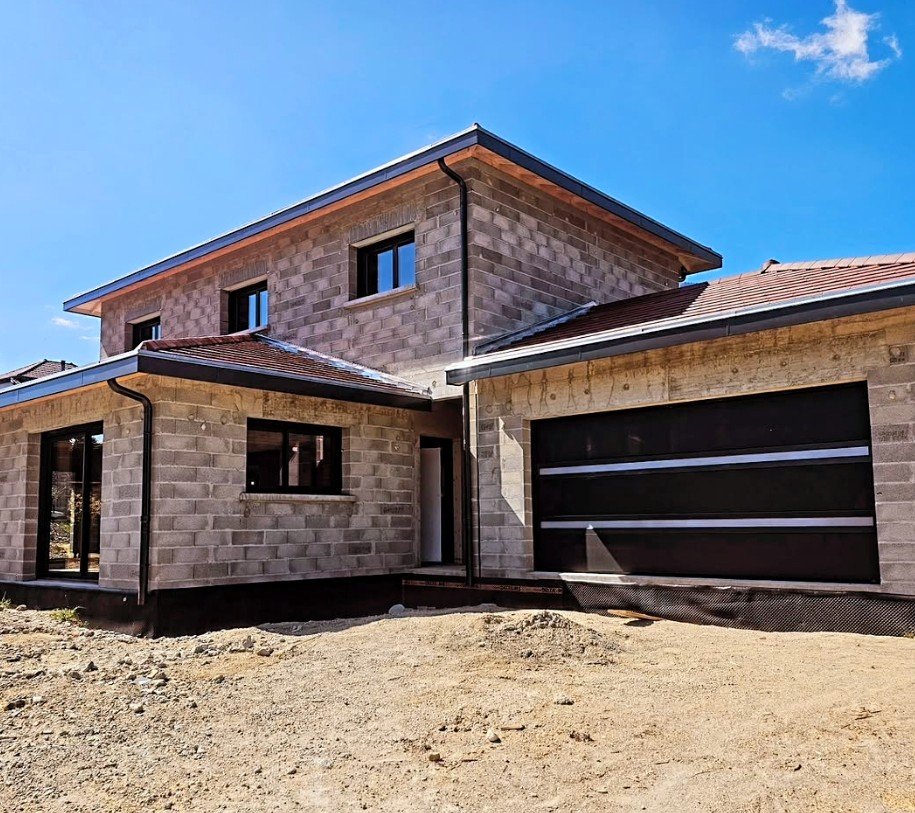 Une maison en construction avec un mur en briques et un garage noir, sous un ciel bleu, sans végétation dans l'avant de la maison.