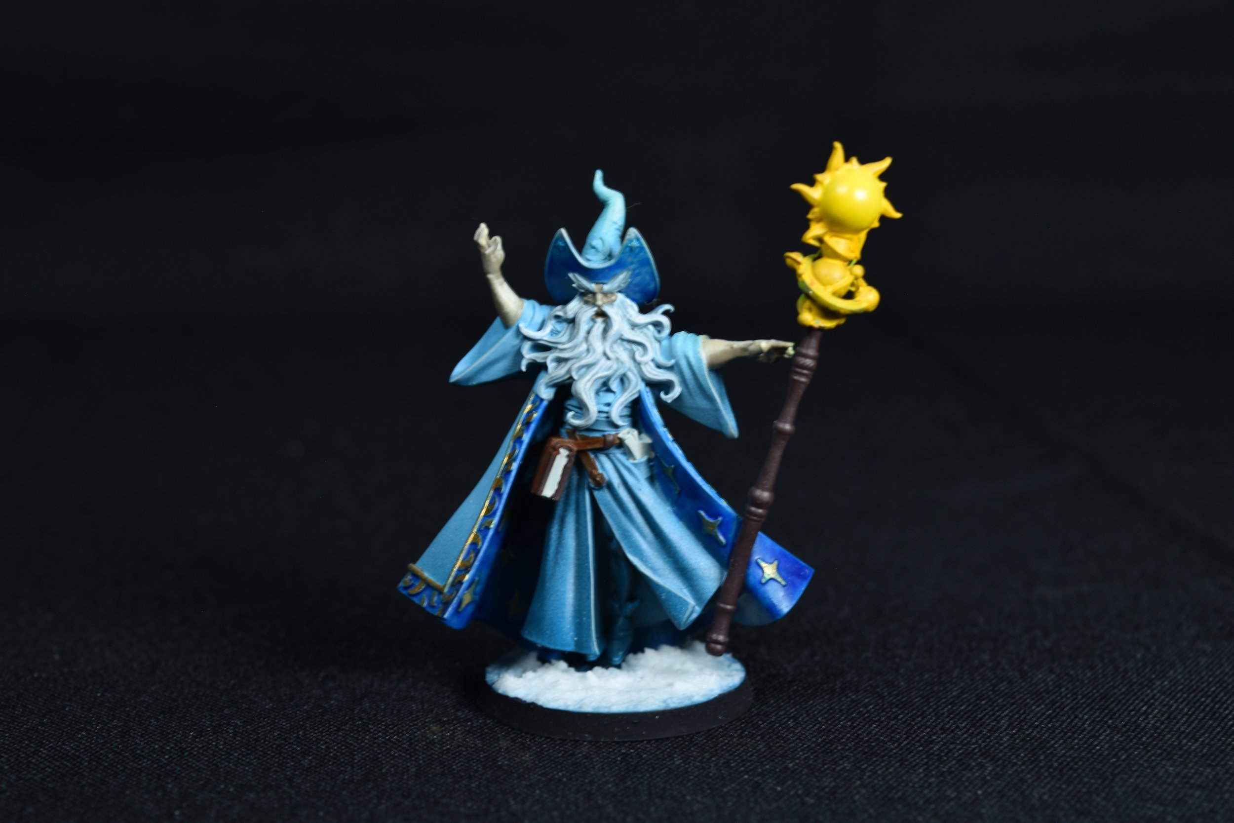 A miniature figurine of a wizard dressed in blue robes and hat, holding a staff with a yellow sun-like ornament, set against a black background.