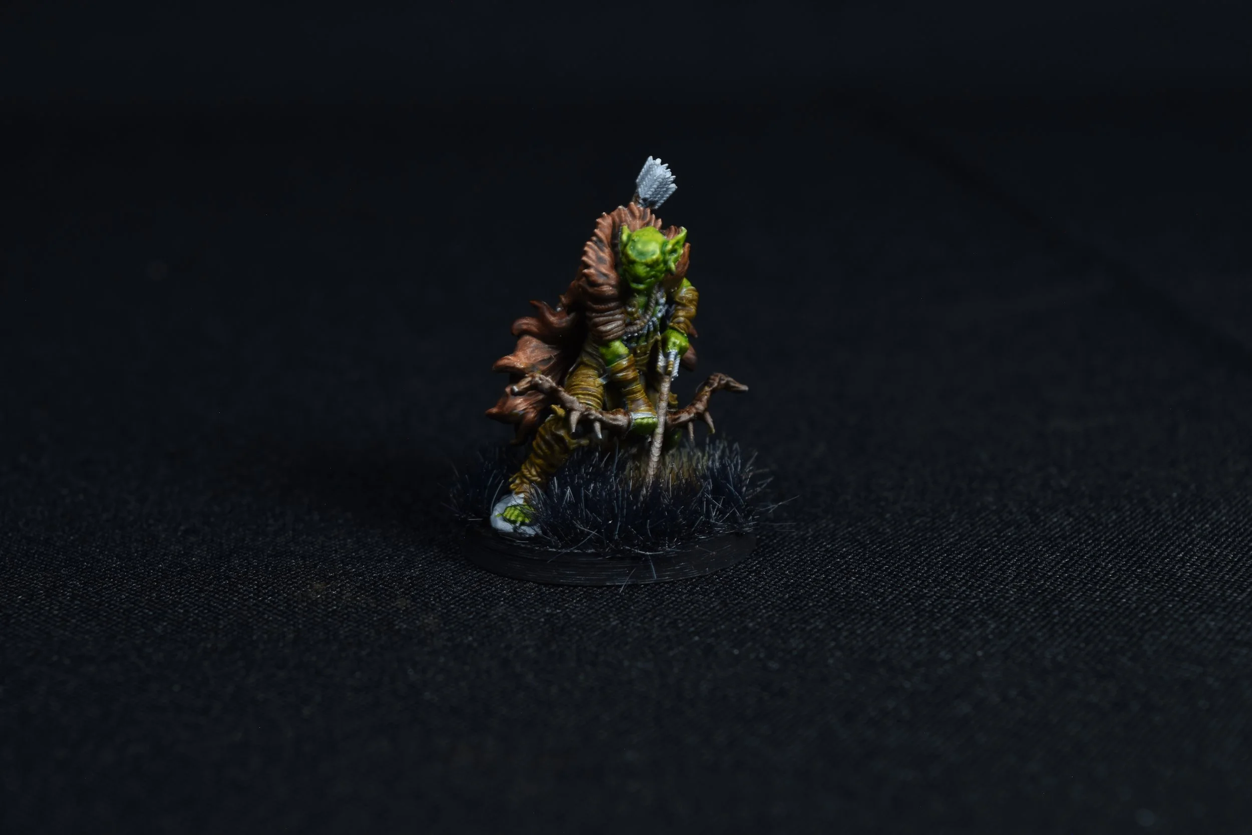 A miniature figure of an orc or goblin warrior holding a sword, wearing armor, with green skin, and a gray feathered crest on its head, standing on a round black base with small tufts of grass, against a dark background.