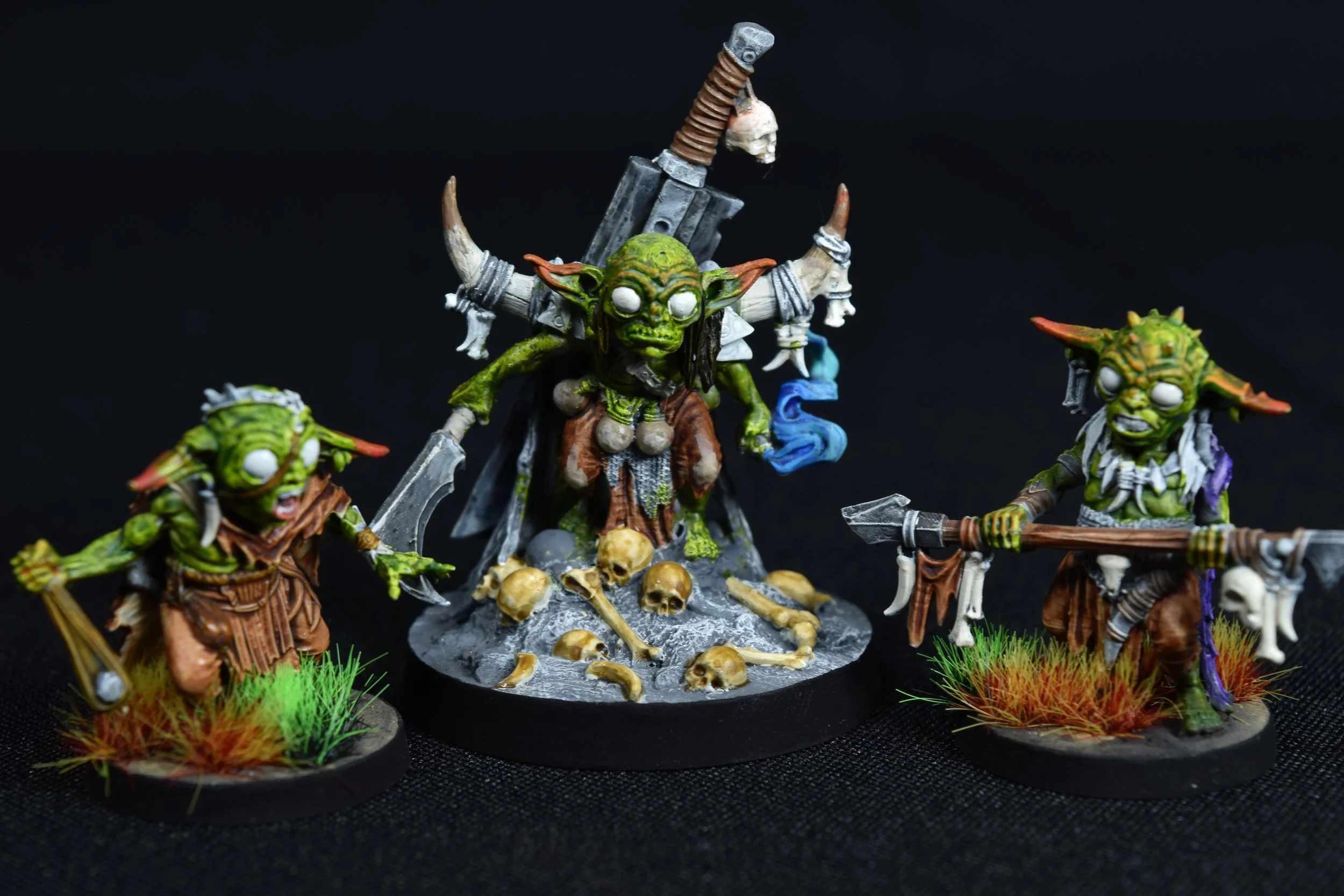 Miniature fantasy orcs with green skin and tusks, carrying weapons, standing around a base with skulls and bones against a black background.