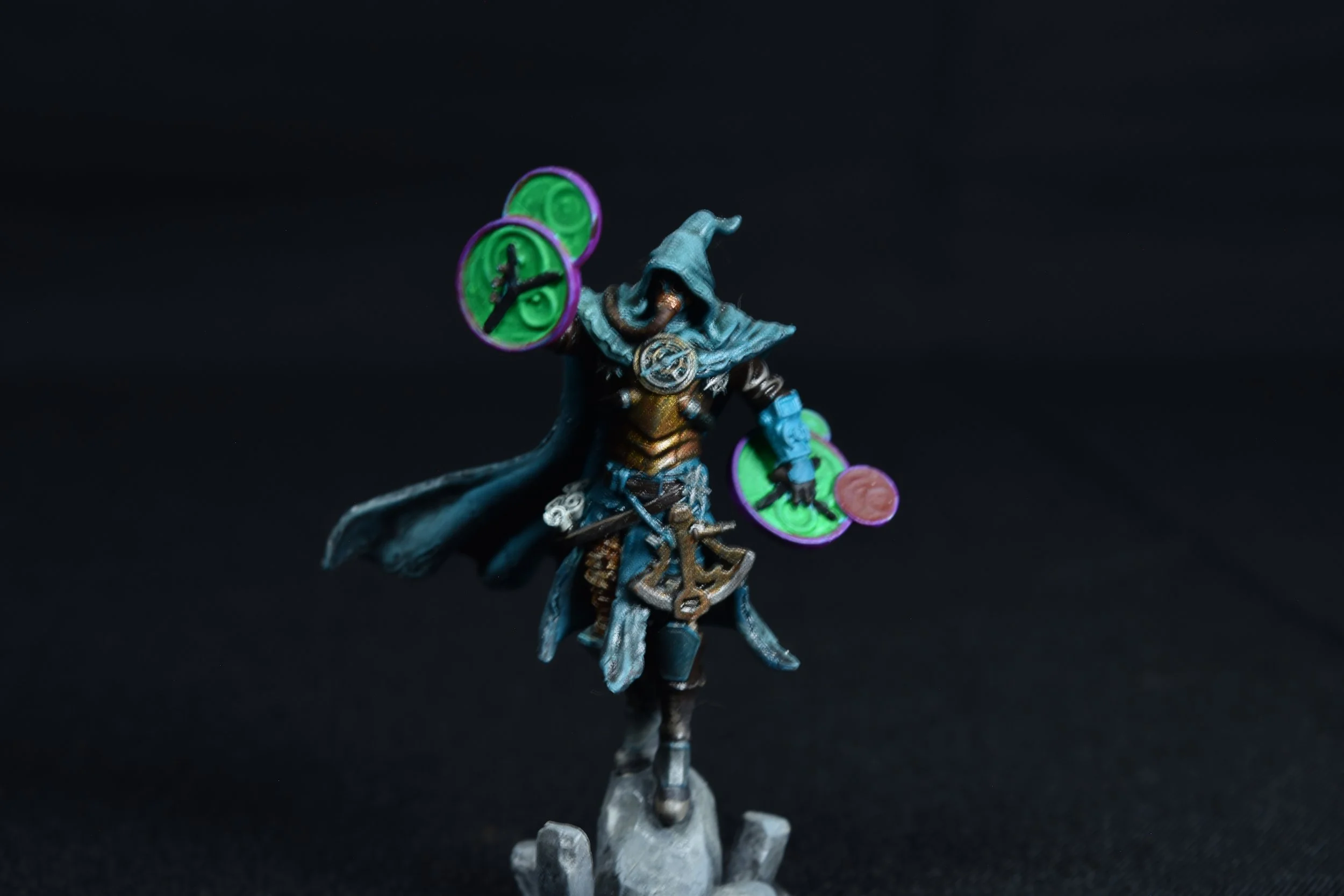 Miniature figurine of a wizard in armor with a hood, holding glowing green discs, standing on a rocky base.