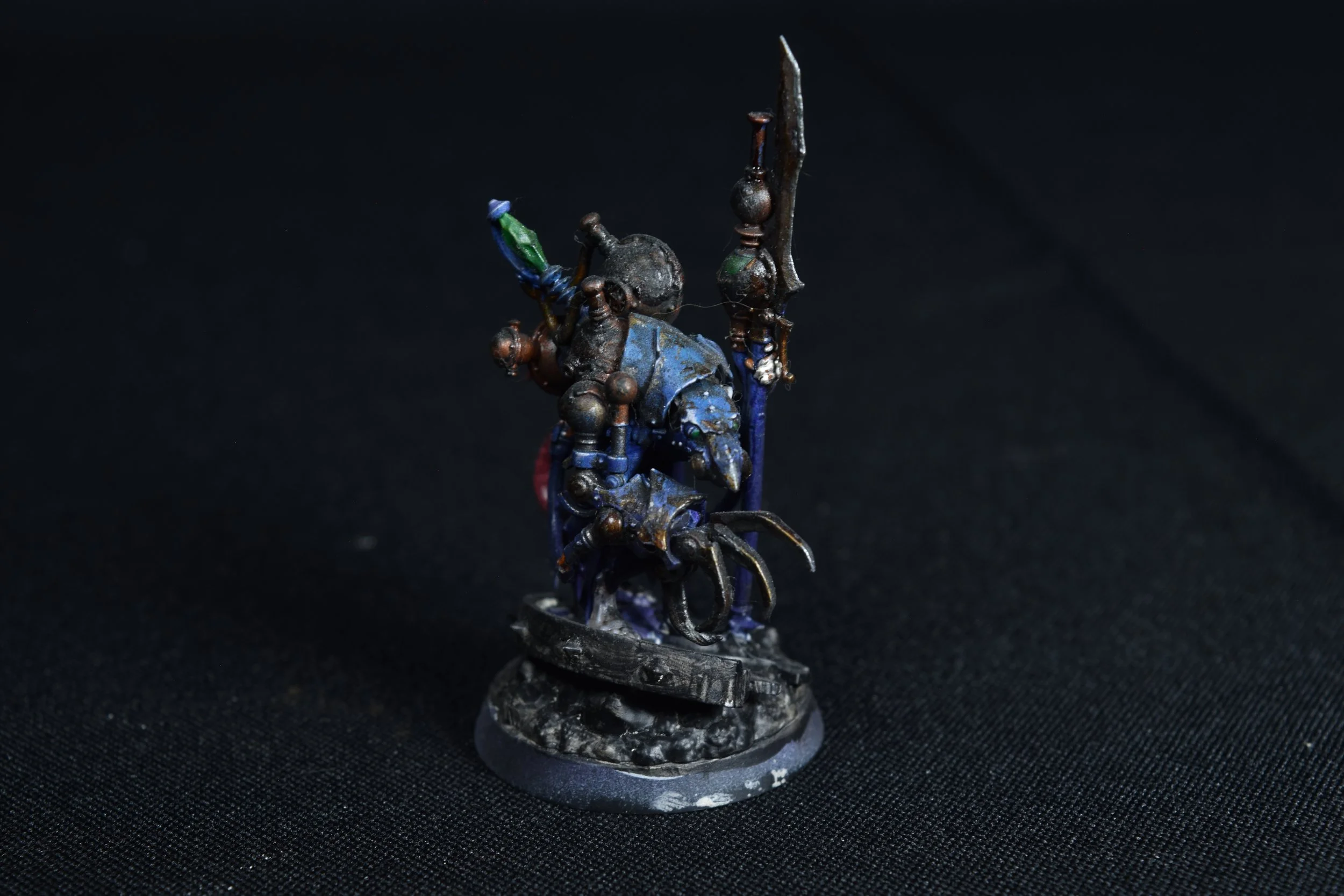 Miniature figurine of a warrior wielding a sword, painted in dark colors with blue accents, standing on a round base with detailed terrain.