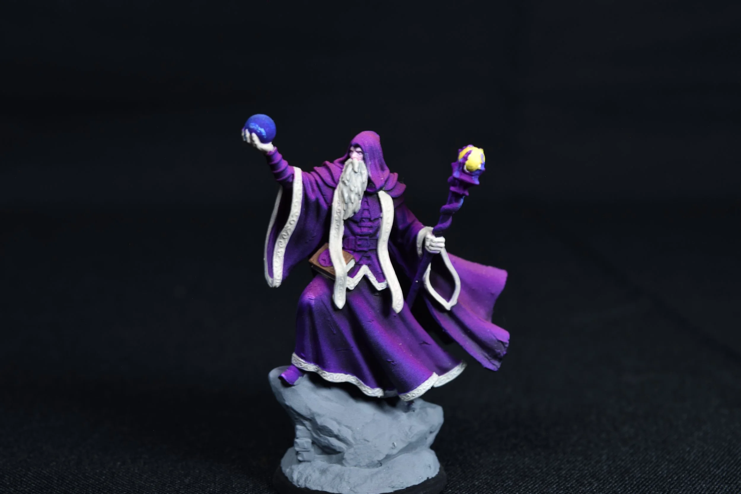 Miniature figurine of a wizard or sorcerer in a purple robe with a hood, holding a purple orb in one hand and a yellow orb with purple accents in the other, standing on a rocky base against a black background.