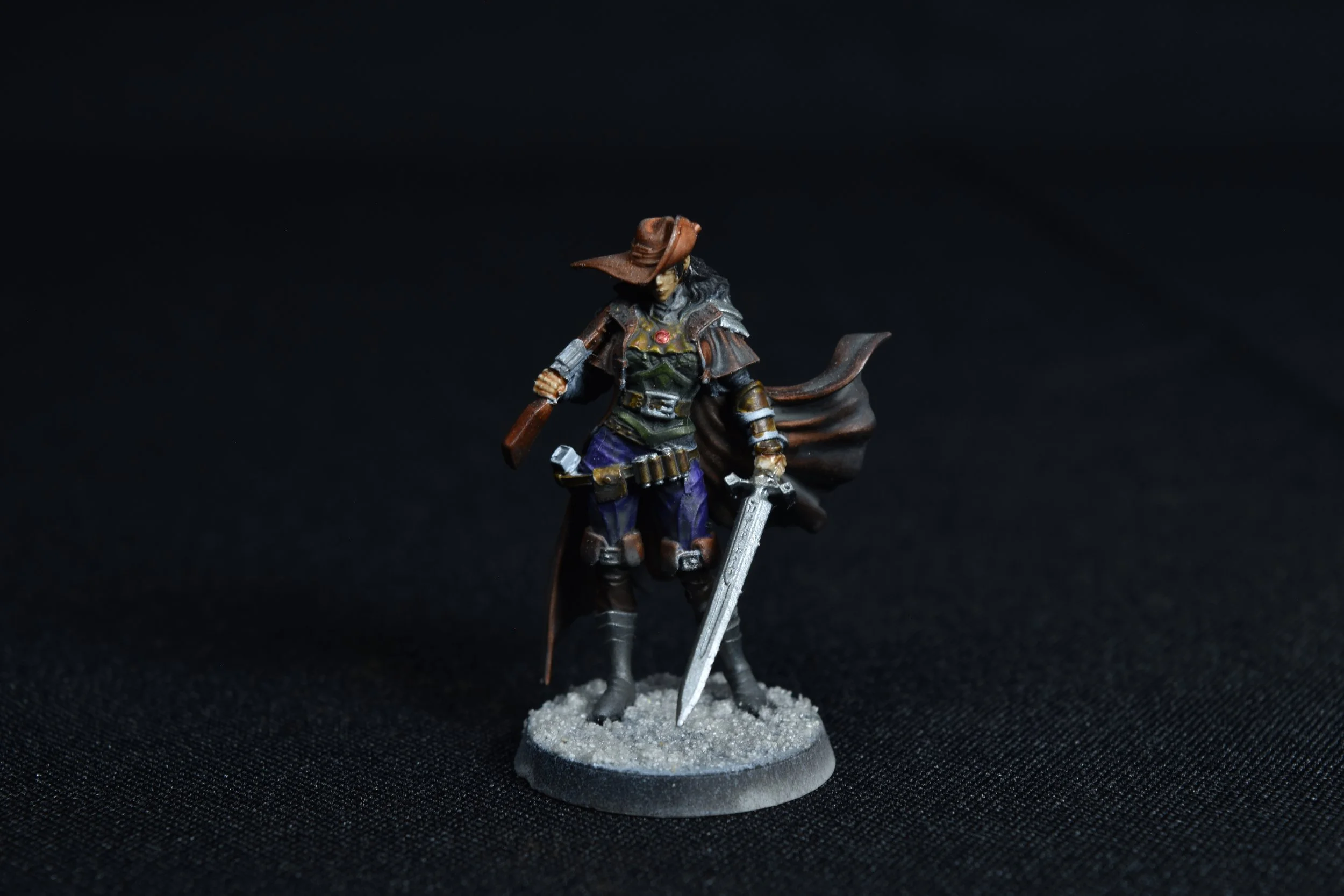 Miniature figurine of a female cowboy with a hat, sword, and crossbow, standing on a round base. The background is black.