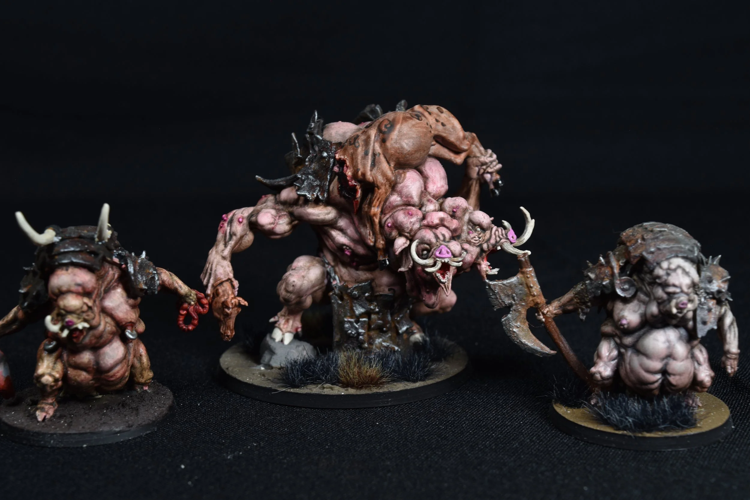 Miniature fantasy creatures with monstrous features, painted in earthy tones, standing on round bases with black textured ground, set against a black background.