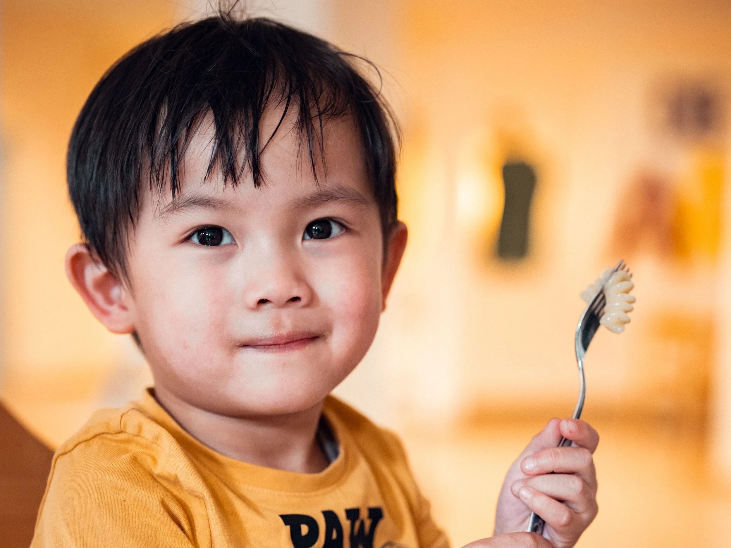 Ein Junge mit dunklem Haar und einem gelben Pullover hält eine Gabel mit ungekochtem Pasta in der Hand. Der Hintergrund ist unscharf und warm beleuchtet.