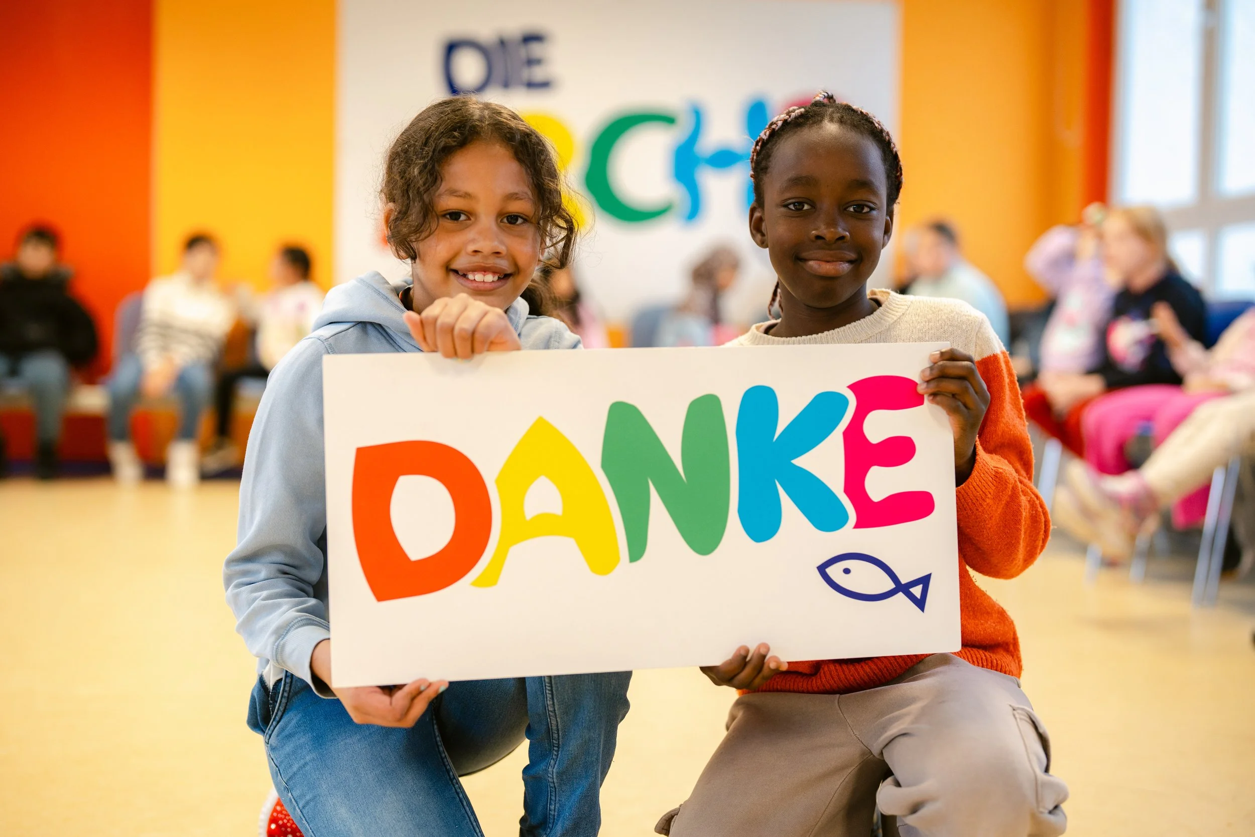 Zwei Kinder halten ein buntes Plakat mit der Aufschrift ‚DANKE‘ in einer Gemeinschaftshalle. Im Hintergrund sitzen weitere Menschen, und die Wände sind in warmen Farben gestrichen.