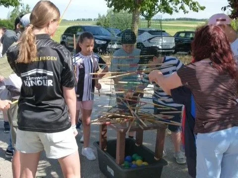 Kinder und Erwachsene spielen ein Holzspiel mit bunten Bällen bei sonnigem Wetter im Freien.