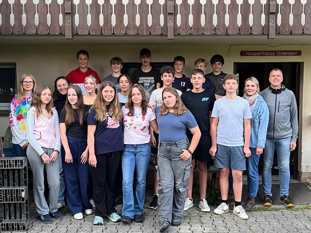 Gruppe von Jugendlichen und zwei Erwachsenen vor einem Gebäude mit Holzveranda, alle lächeln und posieren für das Foto.