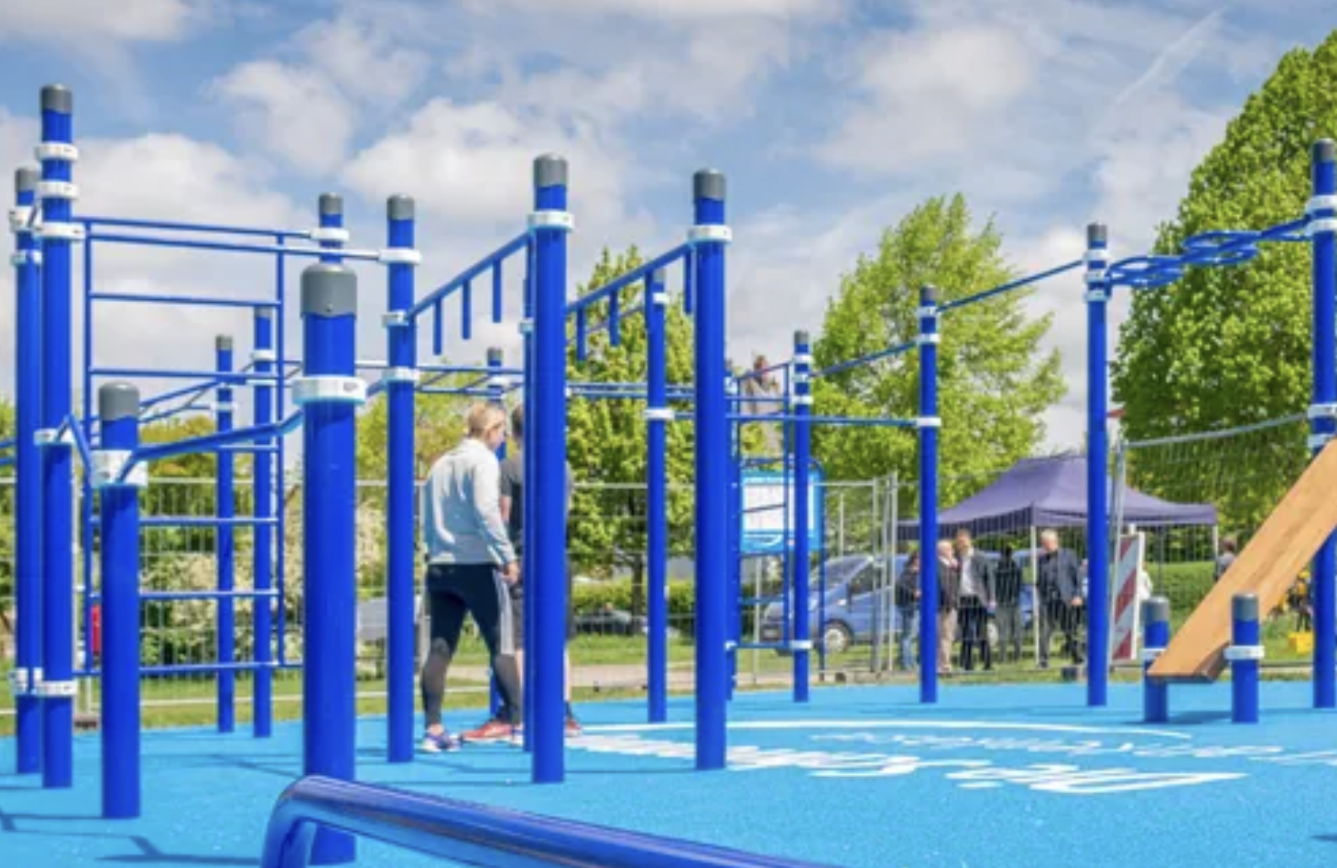 Blaue Kletter- und Schaukelstrukturen in einem Spielplatz mit mehreren Kindern und Erwachsenen, grüne Bäume und ein blauer Himmel mit Wolken im Hintergrund.