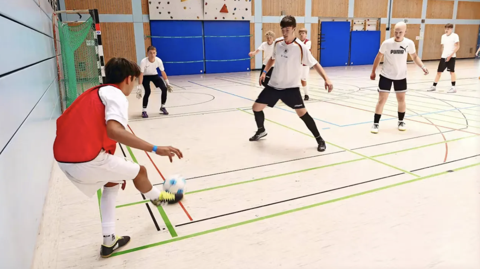 Jugendliche spielen Indoor-Fußball in einem Sporthalle, einer schießt den Ball, während die anderen verteidigen.