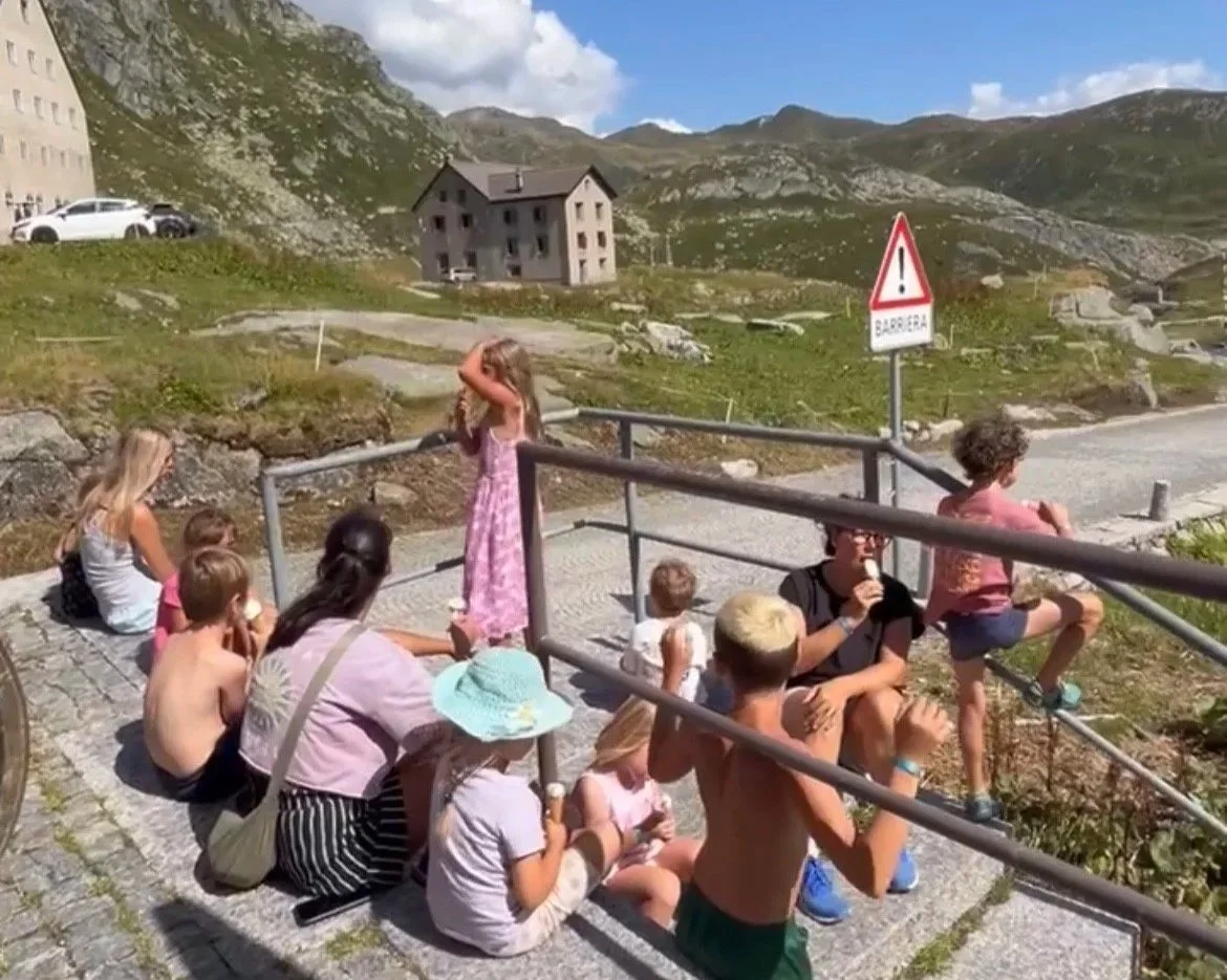 Kinder sitzen und stehen auf einer Terrasse in den Bergen, einige essen Eis. Es gibt eine Warnschilder, das eine Barriere anzeigt, mit grünen, schroffen Hügeln im Hintergrund.
