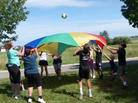 Kinder spielen mit einem großen bunten Fallschirm im Freien, während sie einen Ball darüber halten.