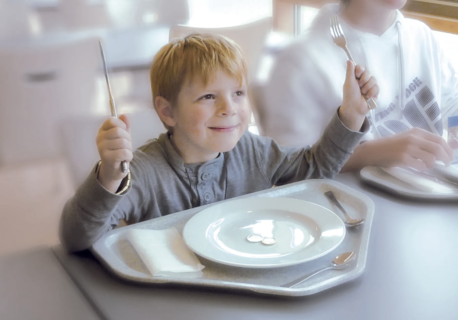 Ein Junge freut sich an einem gedeckten Tisch im Restaurant, hält Messer und Gabel in den Händen, sitzt vor einem leeren Teller.