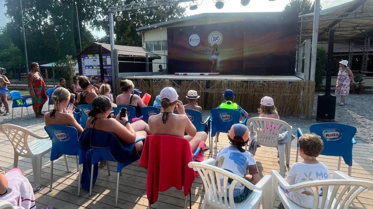 Gruppe von Kindern und Erwachsenen sitzt im Freien und schaut auf eine Bühnenperformance bei einem Sommer-Event.