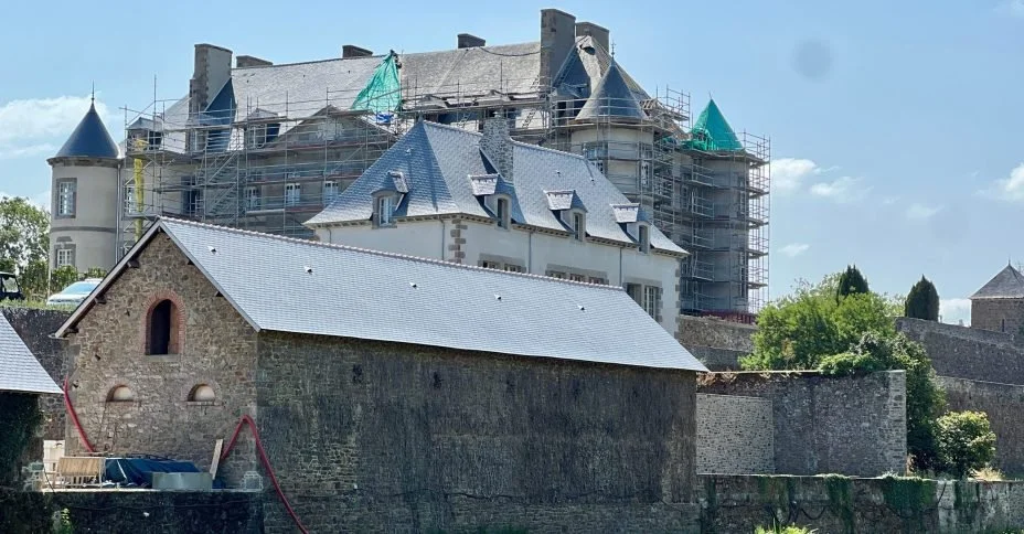 Château en rénovation avec des échafaudages et des toits en ardoise, entouré de murs en pierre et de végétation.