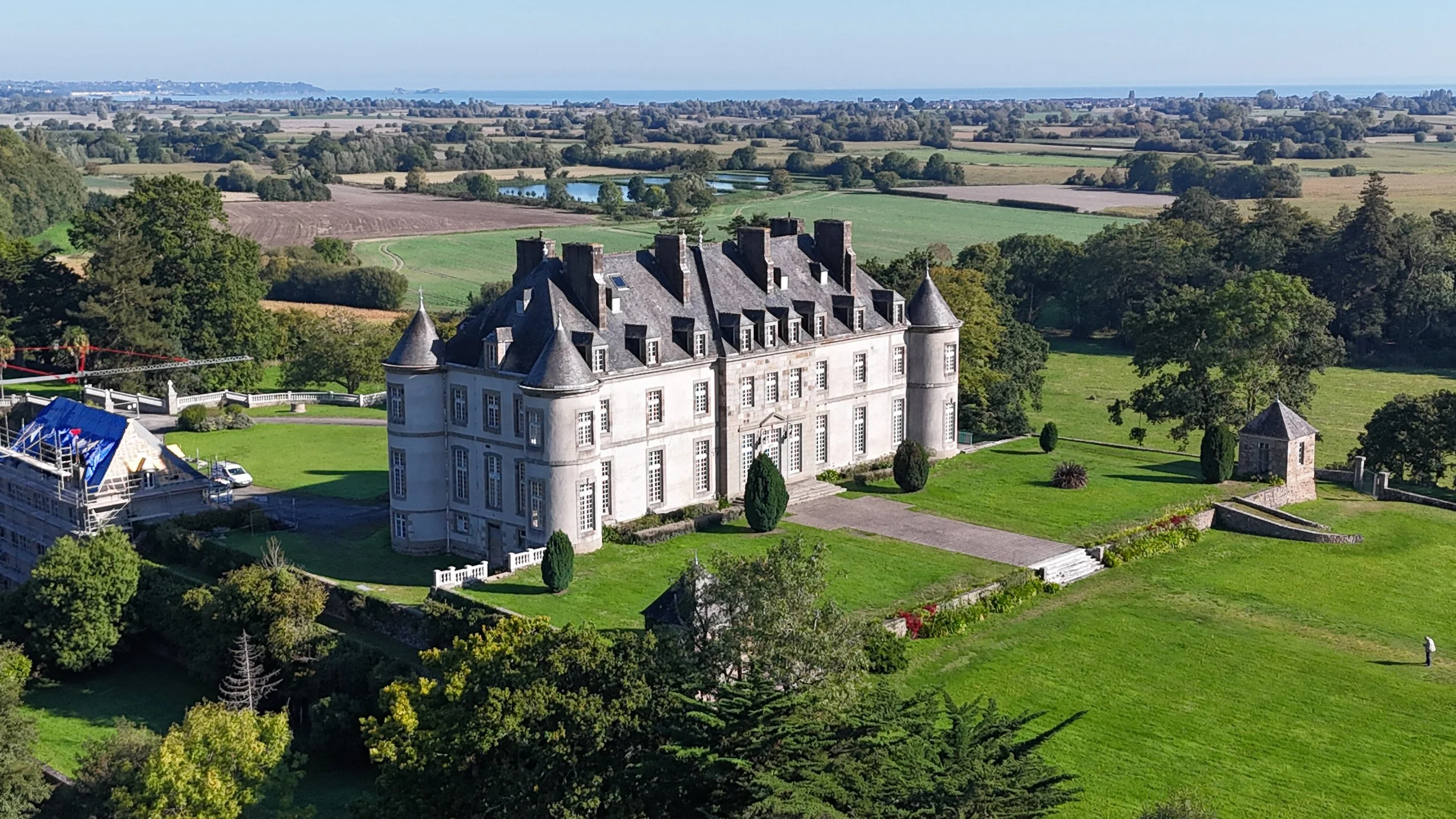 Un château blanc avec des toits pointus et des tourelles, entouré de jardins verts et d'arbres, avec un paysage champêtre s'étendant à l'horizon, incluant des étangs et des champs agricoles.
