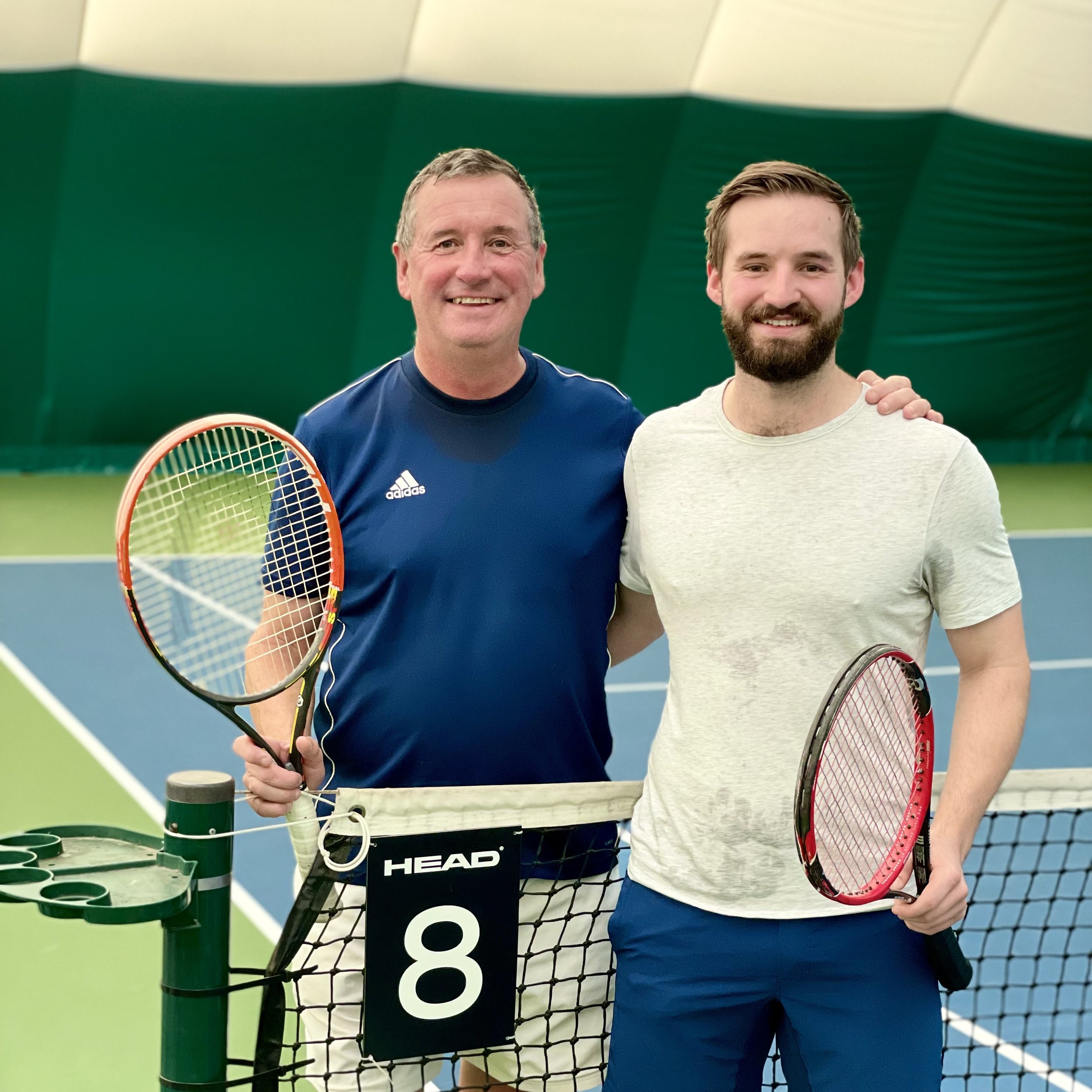 Two male tennis players standing side by side next to a tennis net inside a green and blue indoor court, holding tennis rackets and smiling.