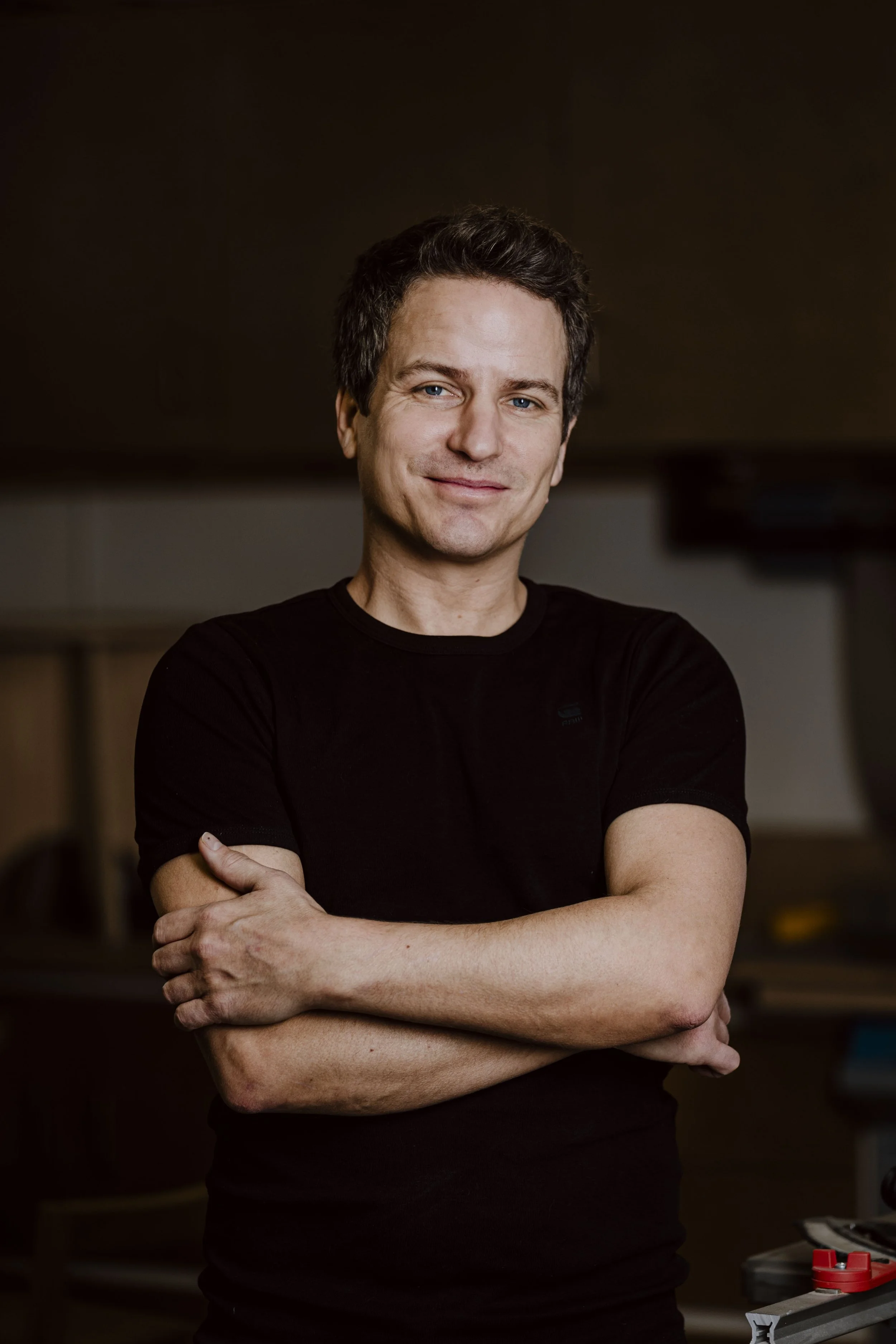 Ein junger Mann mit dunklem Haar, blauen Augen und heller Haut trägt ein schwarzes T-Shirt. Er steht mit verschränkten Armen vor einem dunklen Hintergrund in einem Fotostudio.