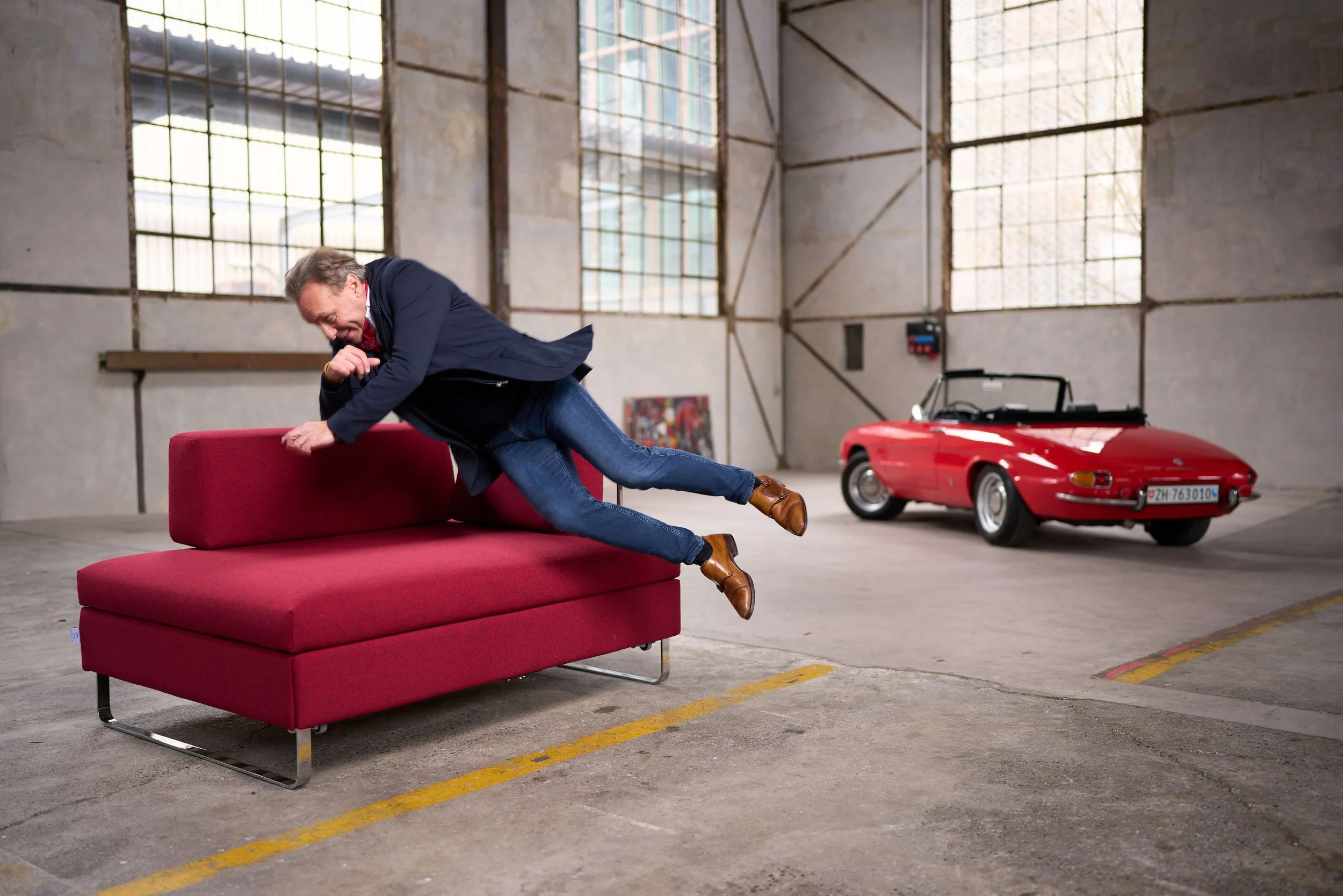 Ein Mann in einem Anzug, der sich auf ein rotes Bettsofa aus Schweizer Produktion stürtz. Das Bettsofa mit der Marke BED for LIVING befindet sich in einer Halle mit hohen Fenstern. Im Hintergrund befindet sich ein rotes Oldtimer-Auto.
