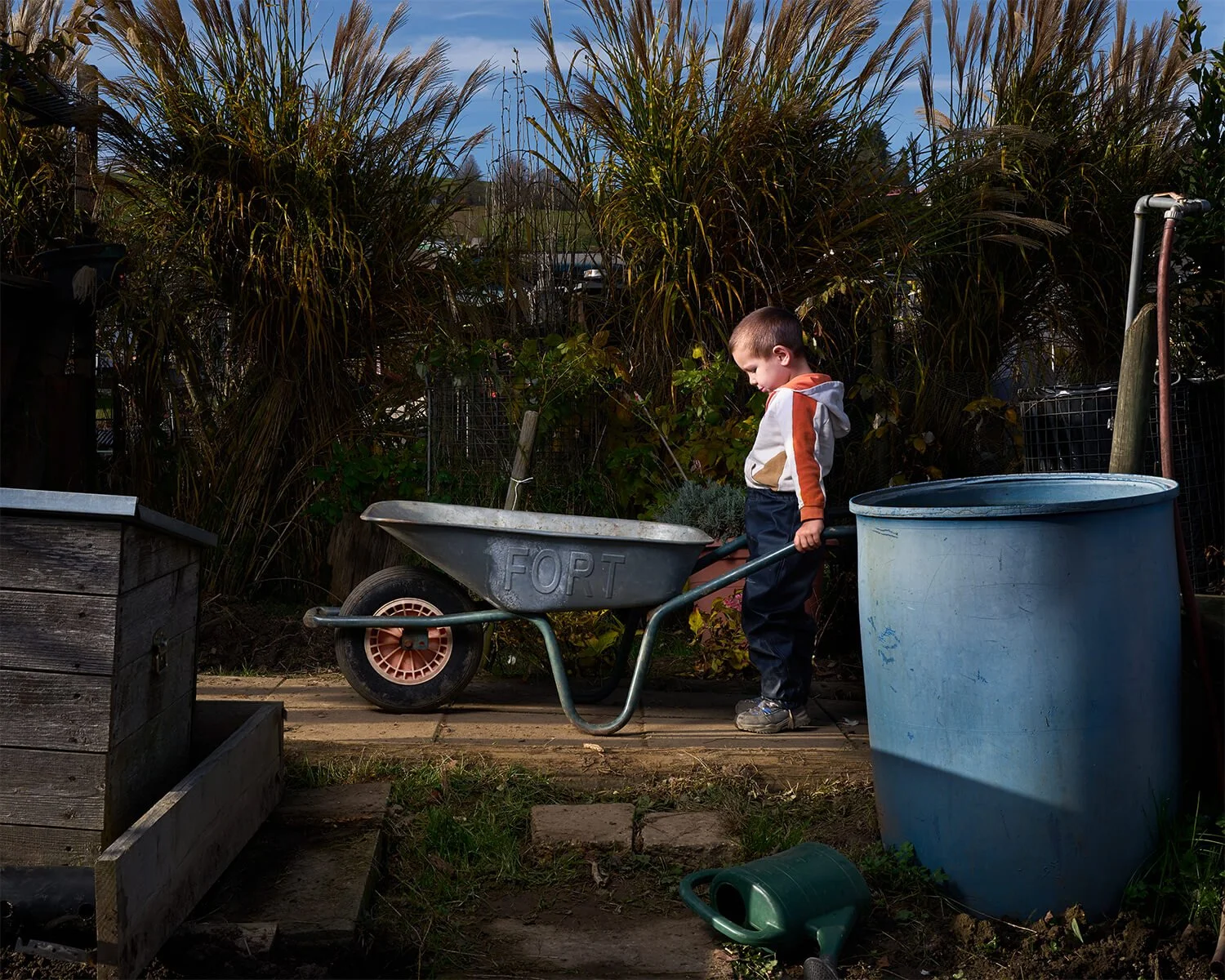 Kontrastreiche Fotografie eines Jungen, der eine Schubkarre mit dem Wort 'FORT' darauf schiebt, steht in einem Garten neben einer blauen Mülltonne. Es ist ein sonniger Tag, und im Hintergrund sind hohe Gräser und ein bewölkter Himmel zu sehen.