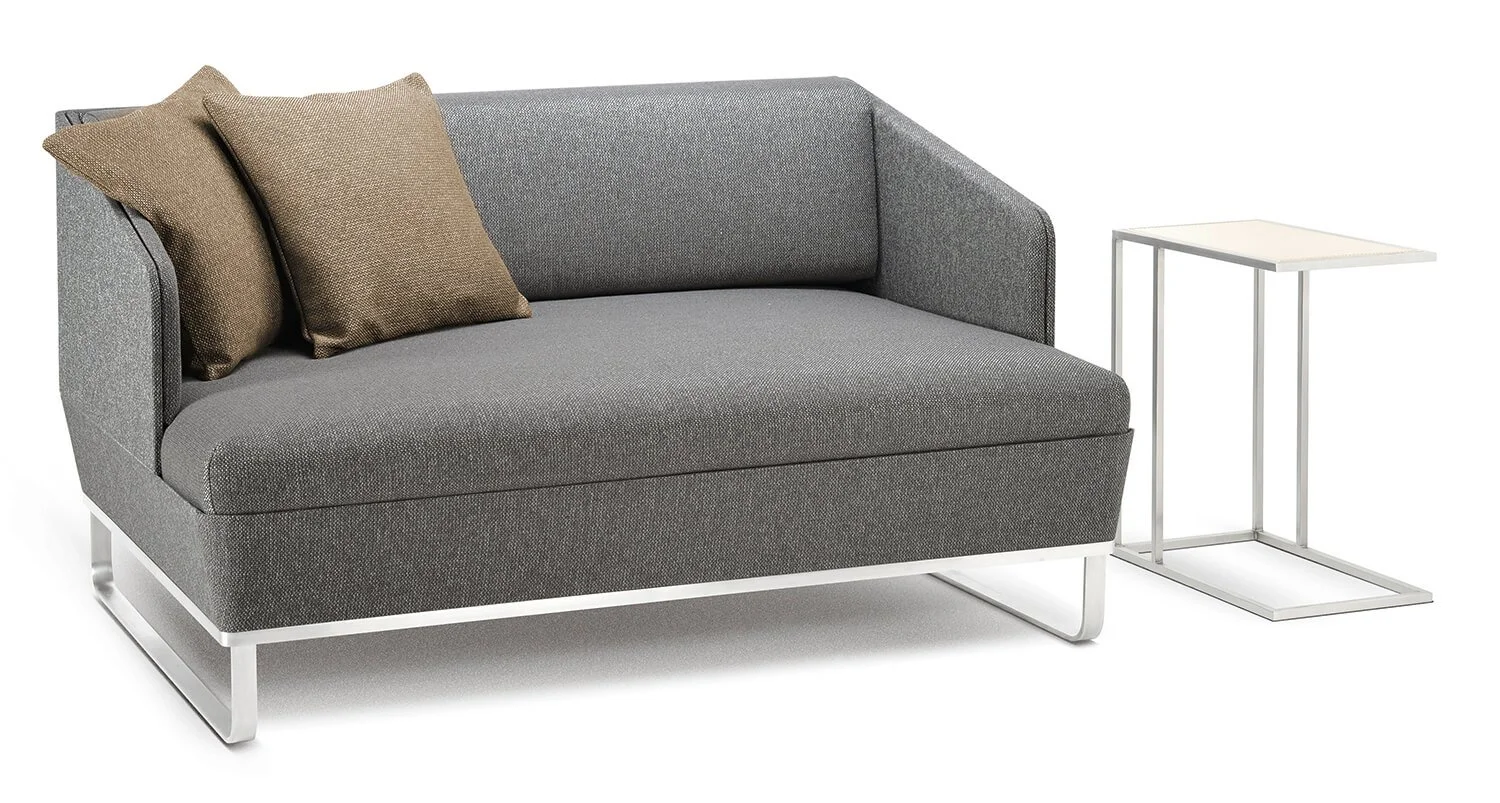 Ein modernes graues Sofa mit zwei braunen Kissen und einem kleinen weißen Beistelltisch daneben.